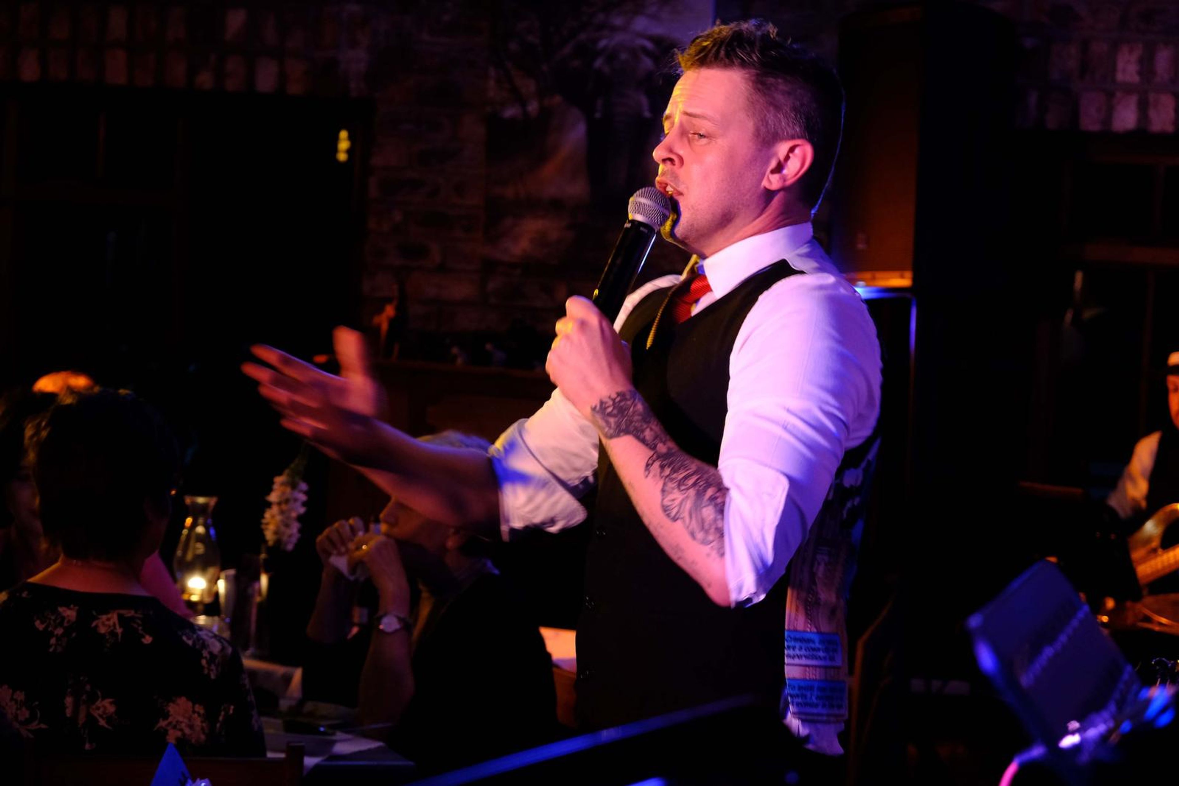 Singer in a waistcoat singing with a hand open to the room