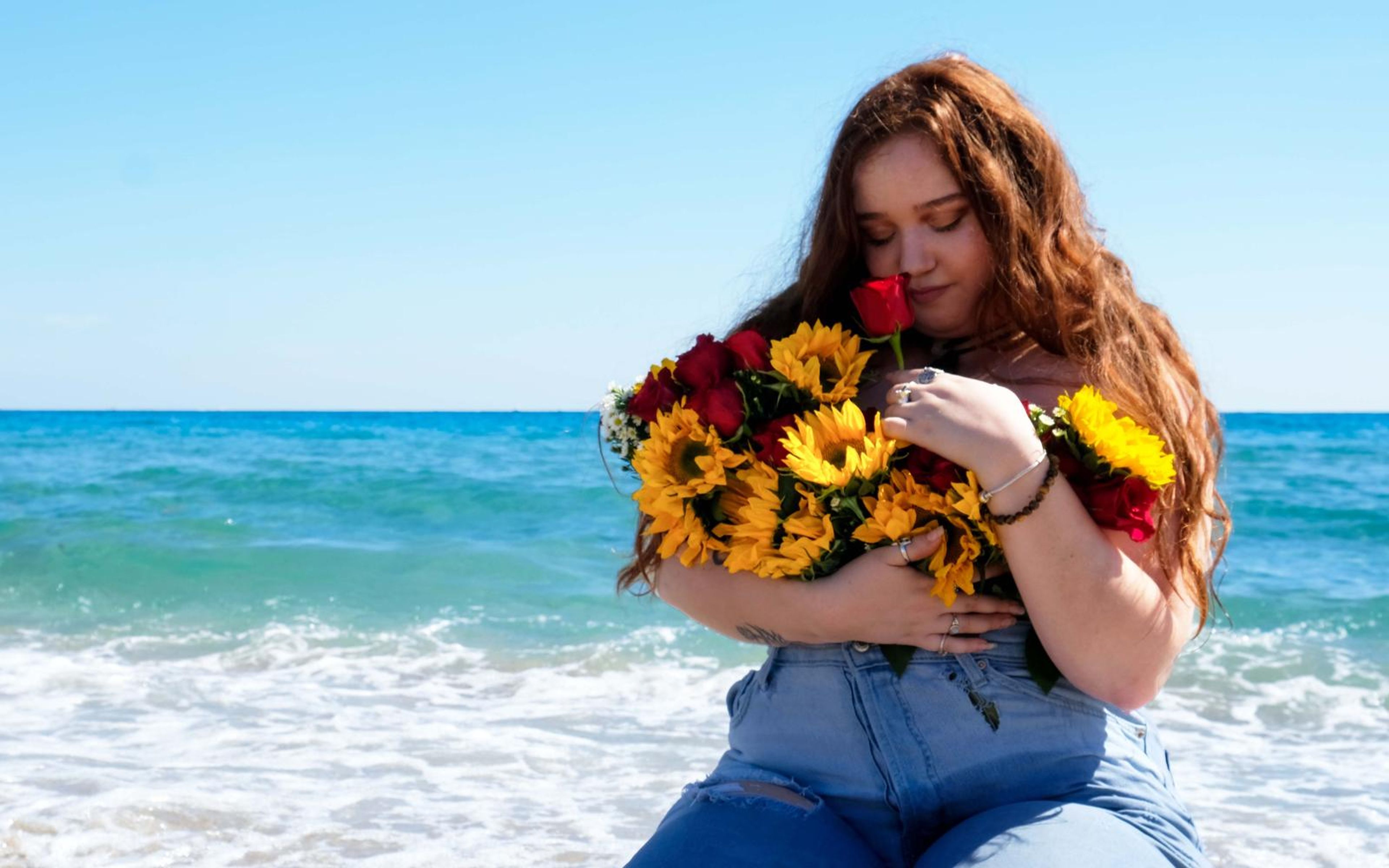 Woman seated at the water's edge with eyes closed, smelling a red rose from a sunflower bouquet held against her chest, ocean behind