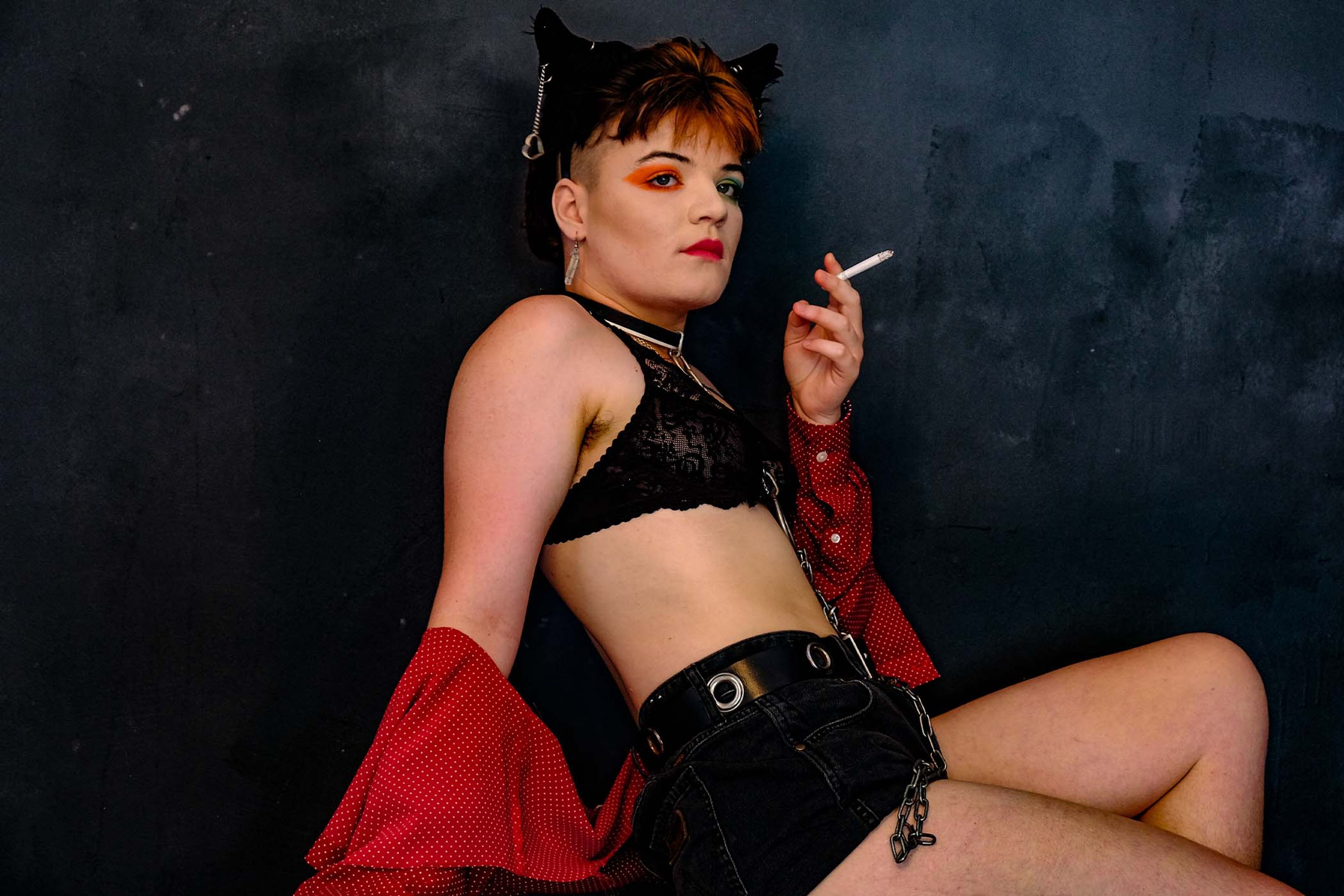 Portrait, subject seated in a lace bra and denim shorts smoking a cigarette, cat-ear headpiece