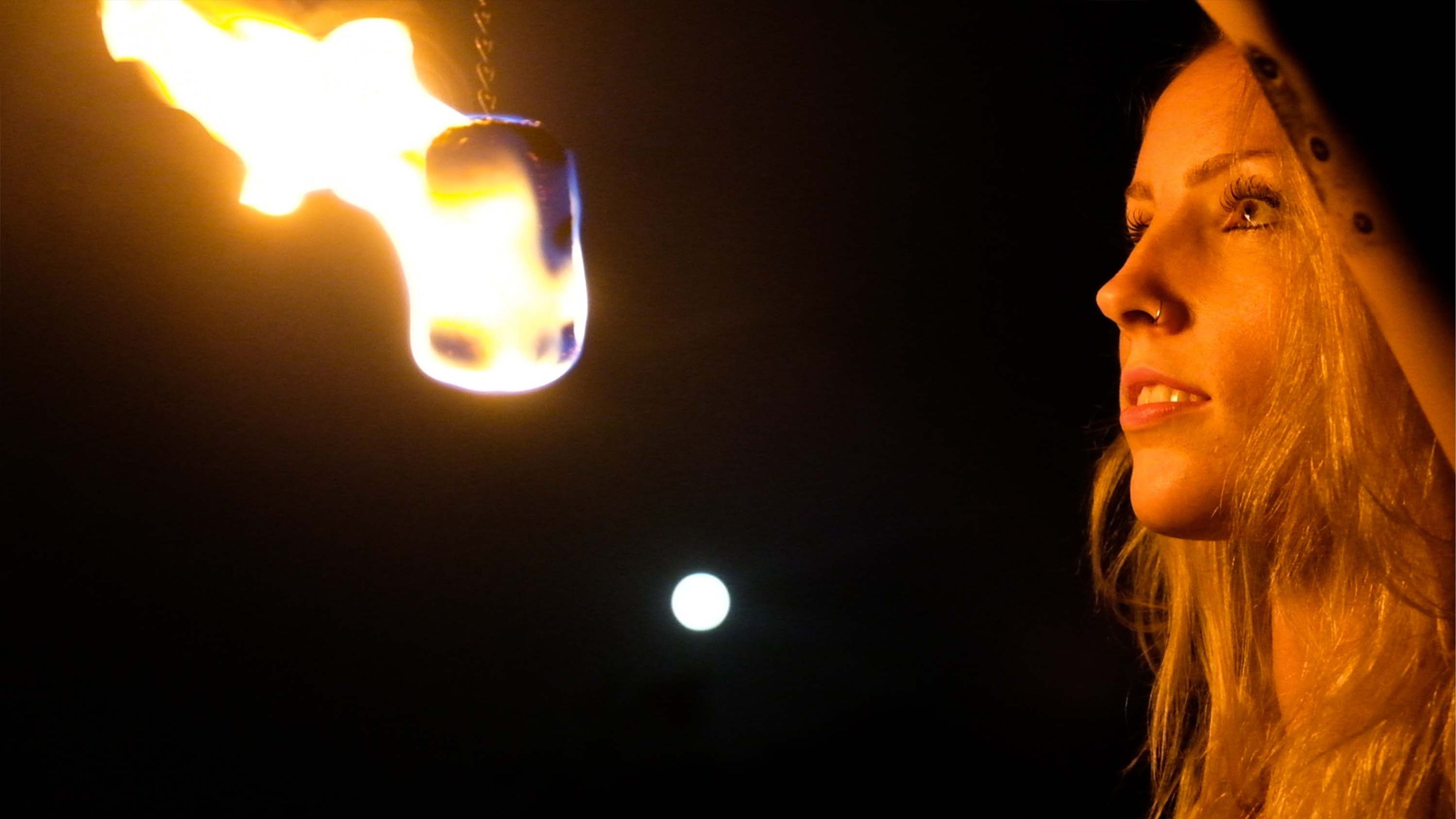 Man with both arms raised overhead and fire poi flaring at each side, dark blue dusk sky behind