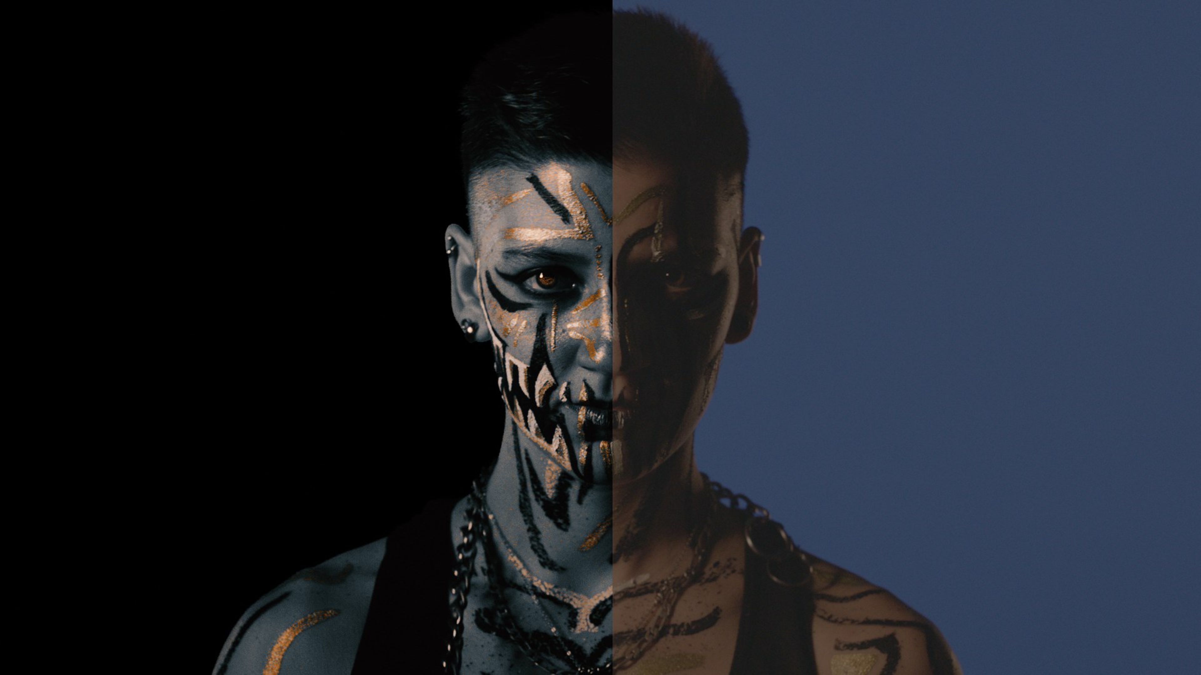 Face-painted portrait with black and gold fang markings, half in shadow against a blue backdrop