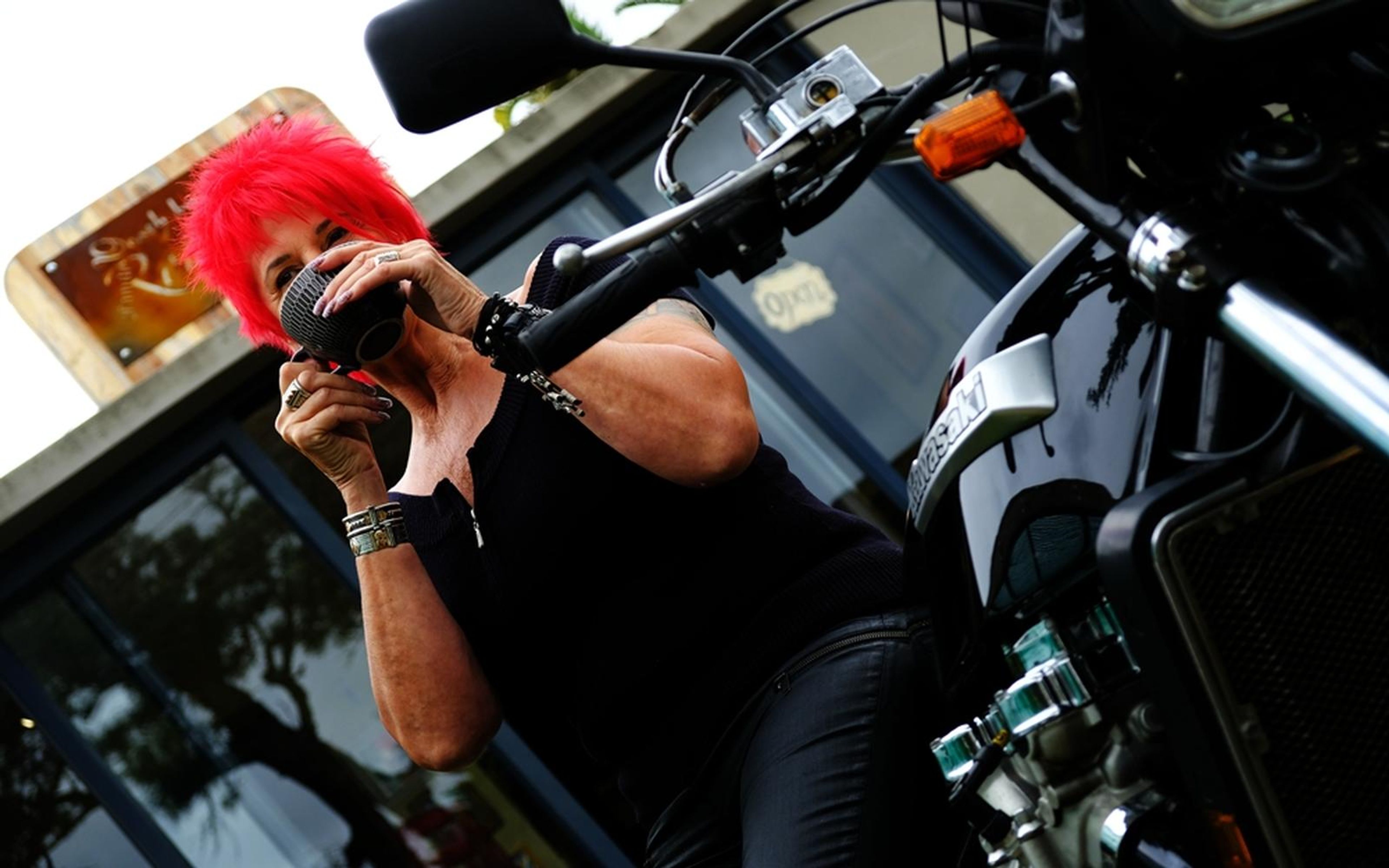 Woman with red hair drinking coffee with both hands, close view over a black motorbike handlebar