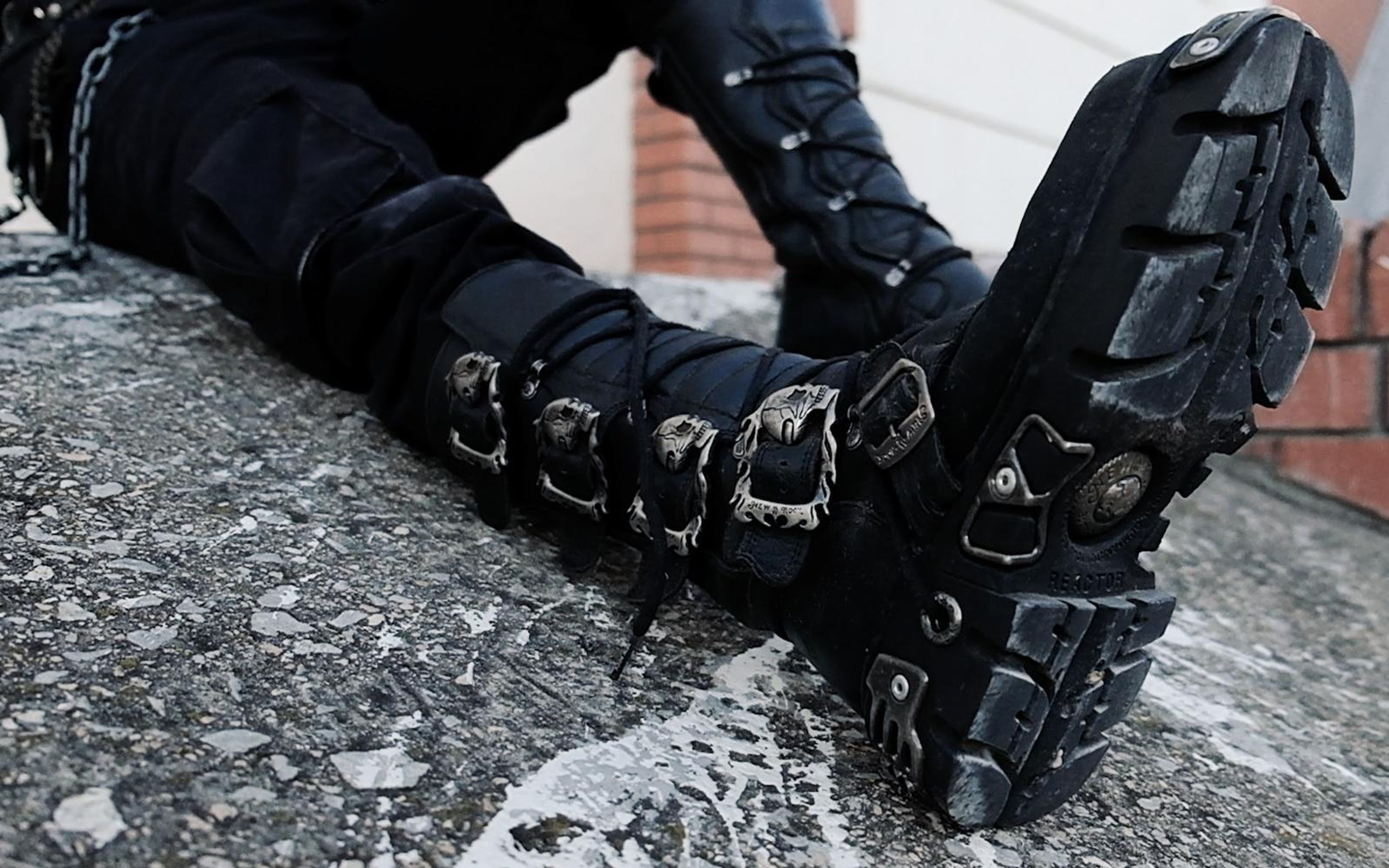 Crossed boots on a concrete ledge with soles and buckles in focus, brick wall and black chained clothing behind