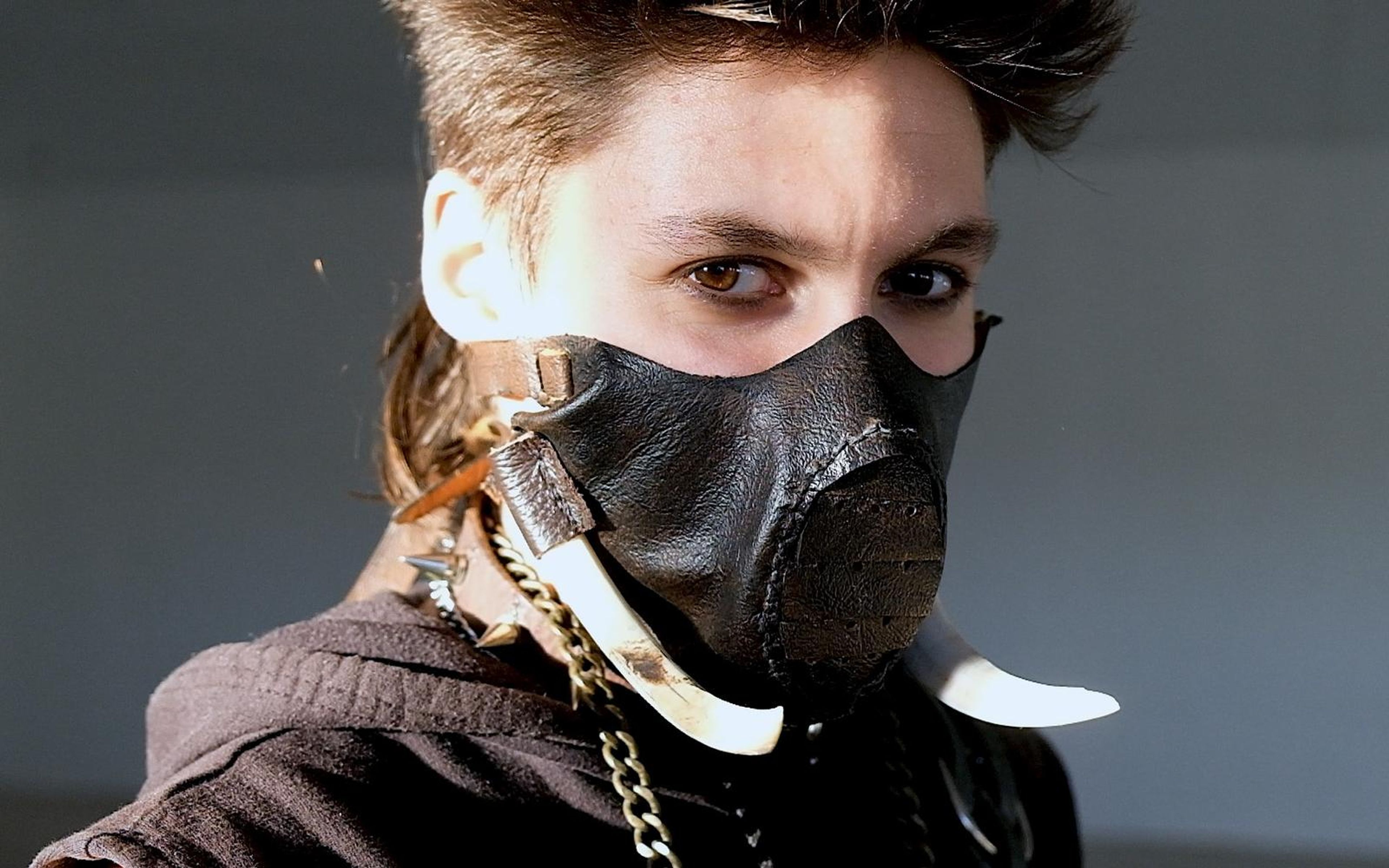 Close frontal view of a masked figure in a studded leather mask with tusk and chain, intense stare, dark backdrop