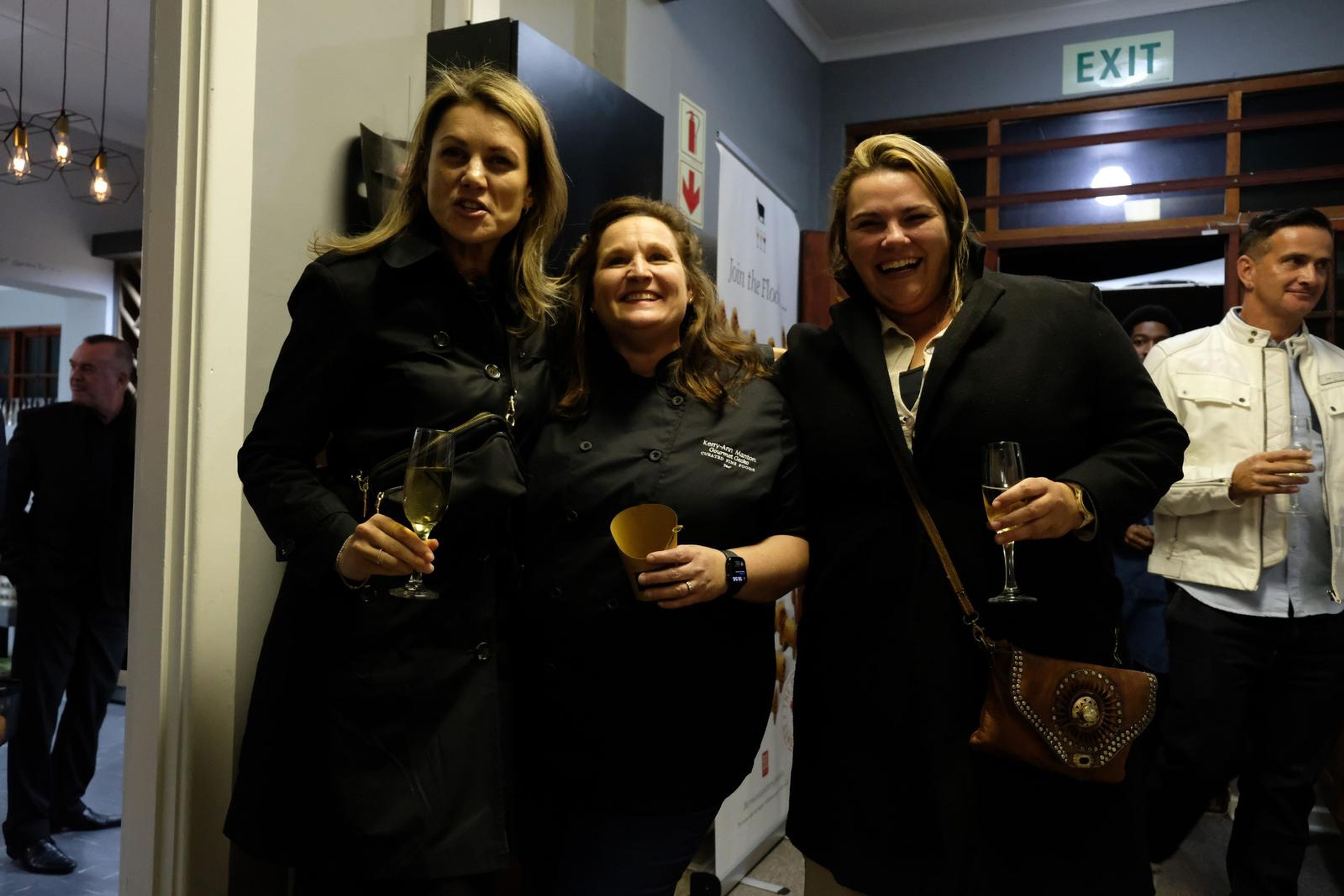Three women in black coats and Chef's Larder shirts smiling together holding drinks