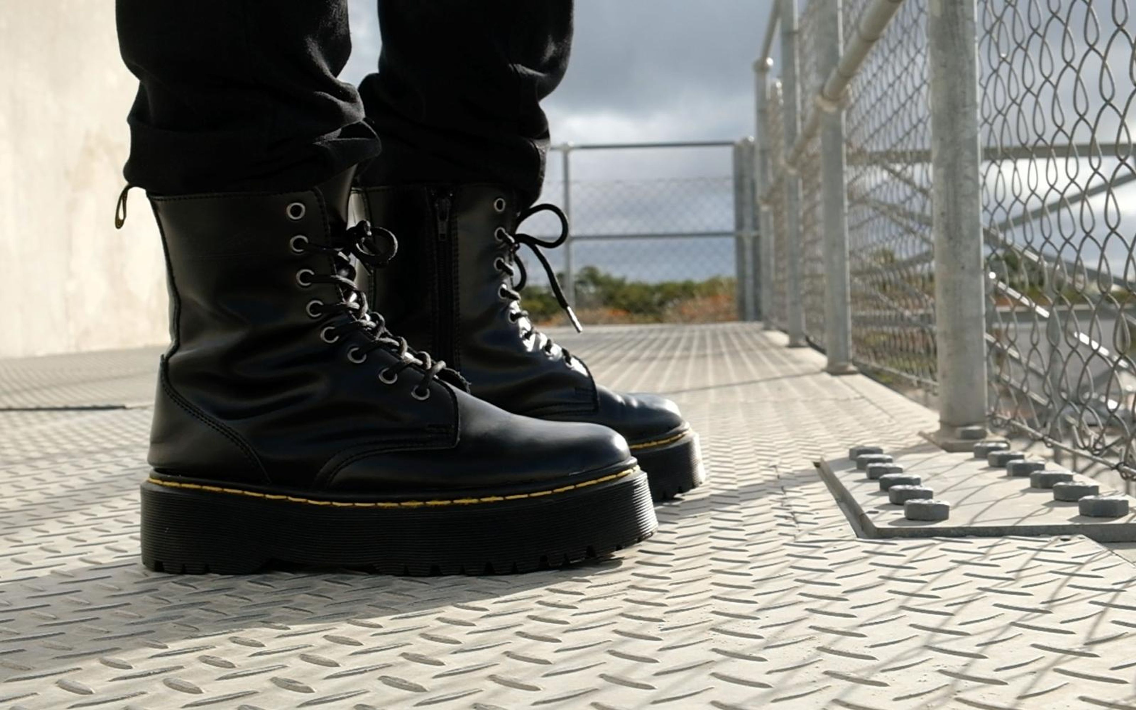 Pair of black platform boots seen from the front on a steel walkway, cloudy sky through the fence