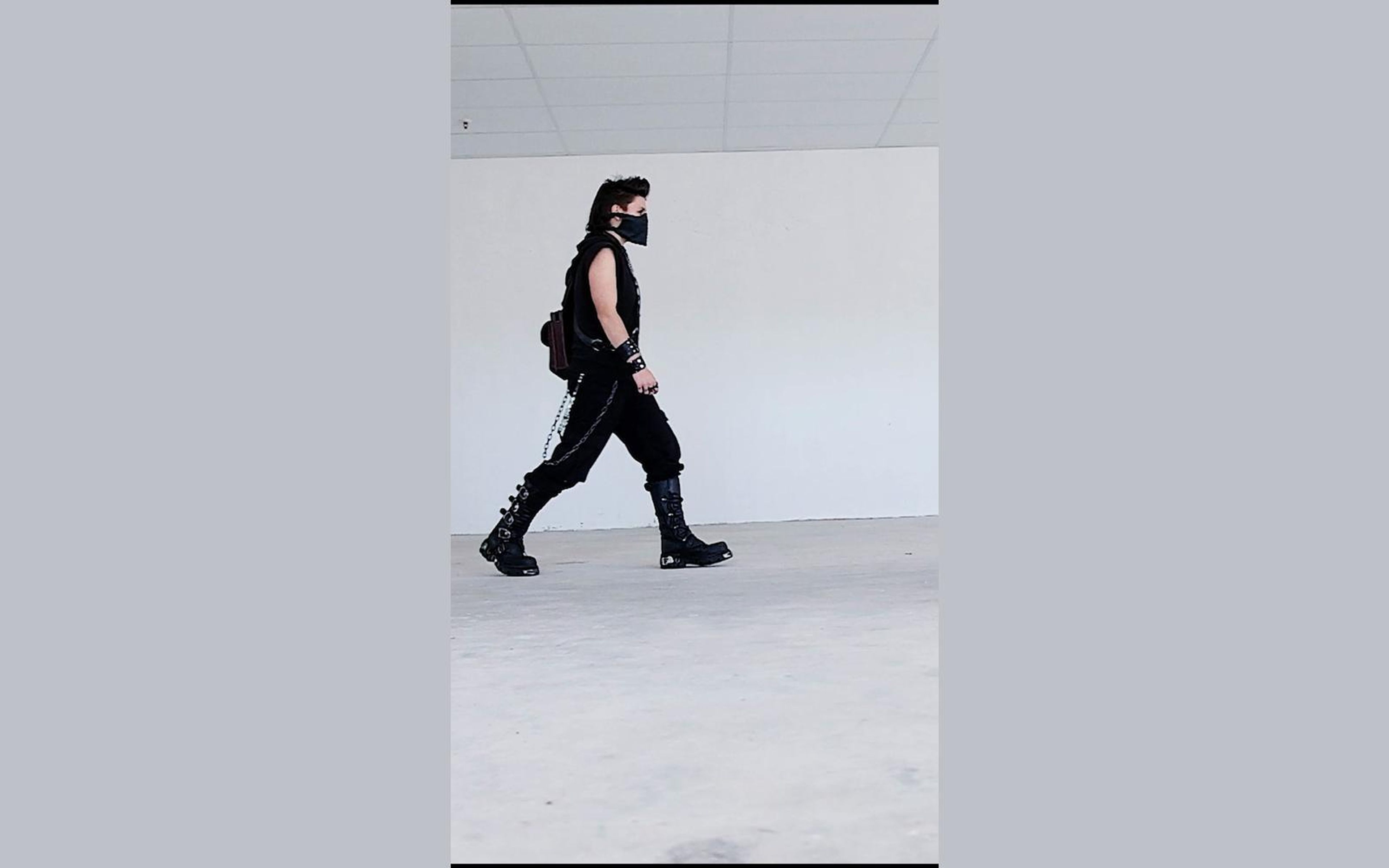 Masked figure mid-stride in profile in New Rock boots across a pale concrete floor, chains swinging at the hip