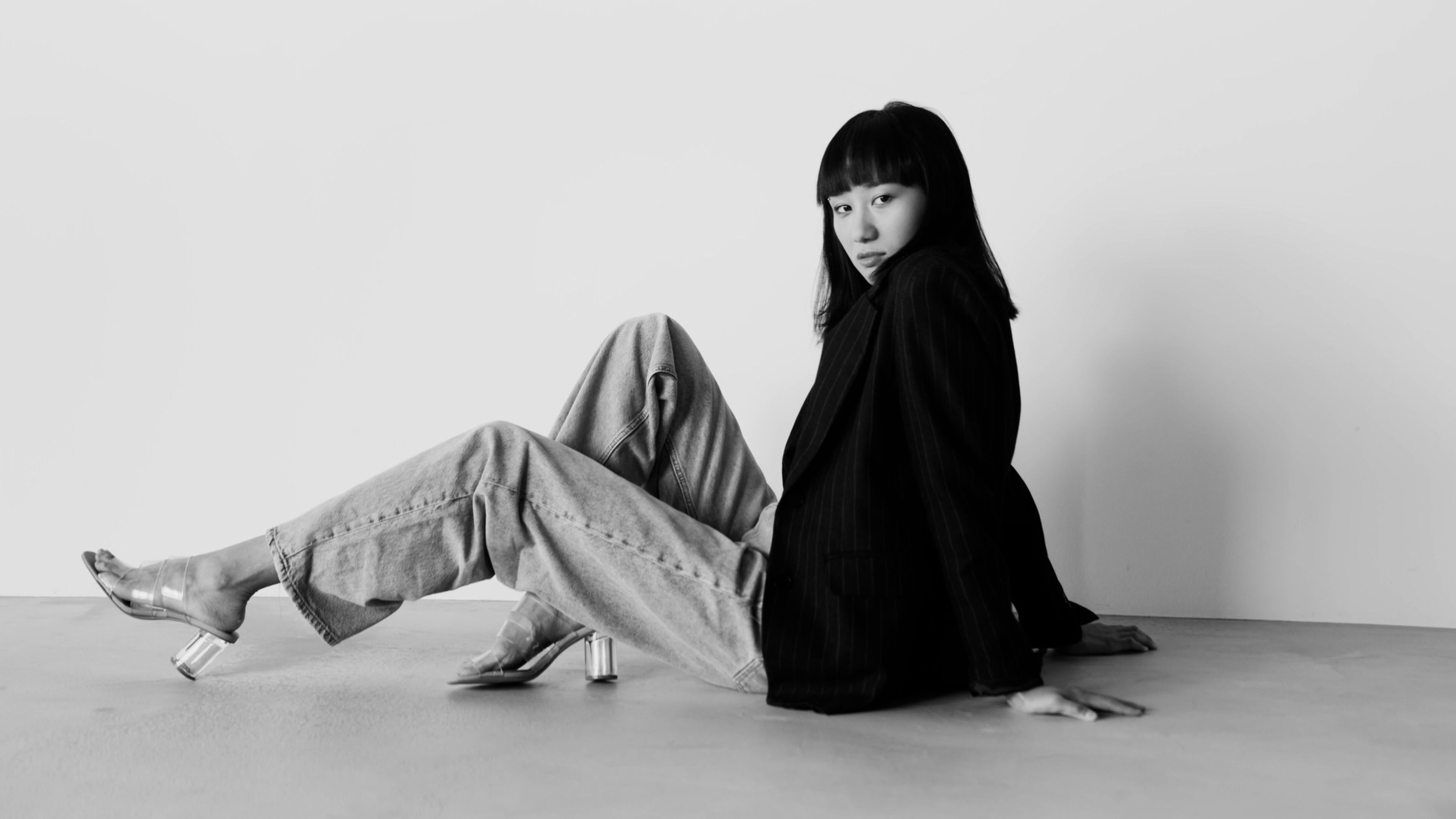 Woman in grey trousers and a black jacket seated on a white studio floor