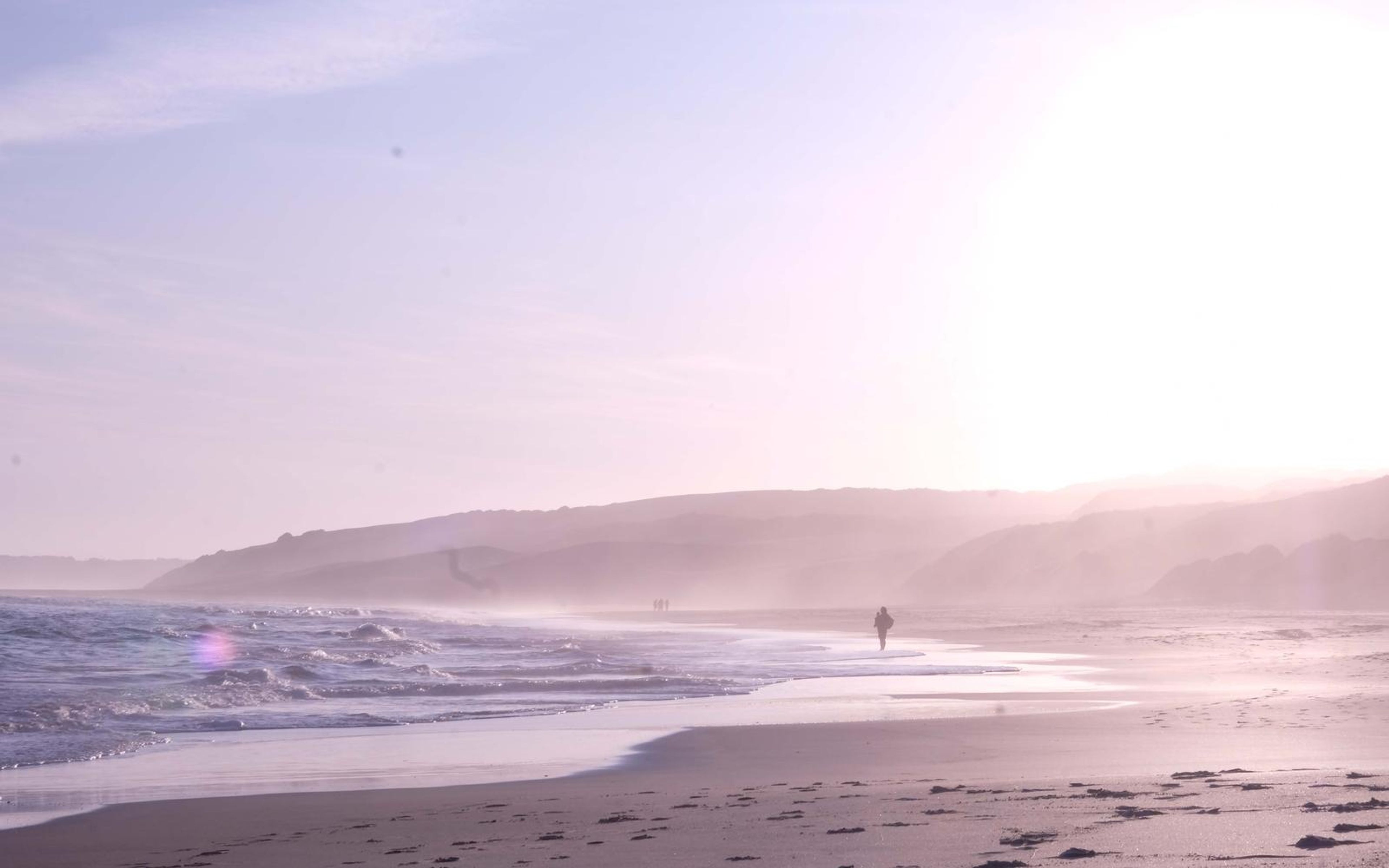 Hazy shoreline with a tiny distant walker, backlit sun and sea mist over the hills