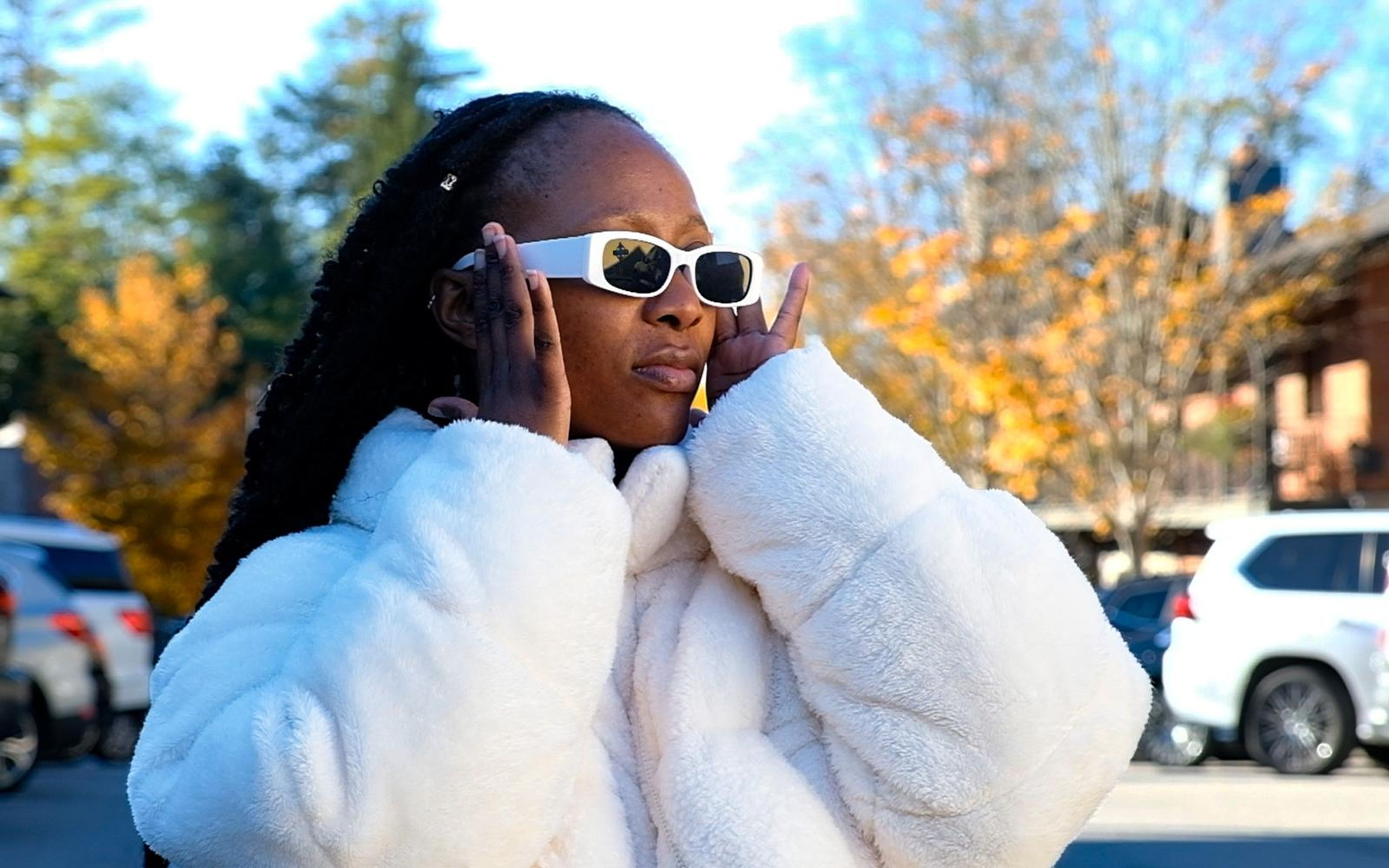 Model holding the arms of her white sunglasses with both hands, puffy fur jacket, warm autumn background