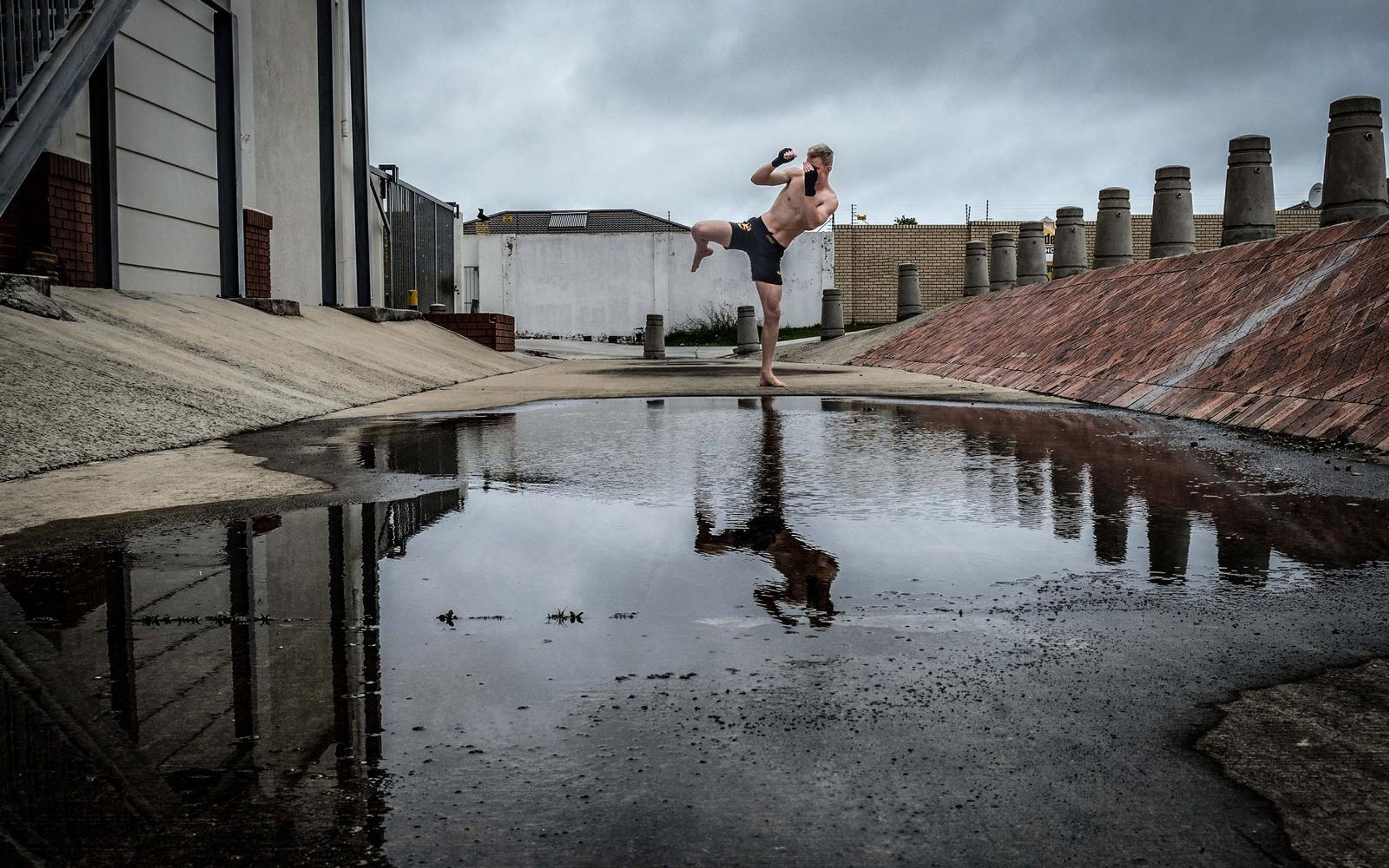 Man in a high kick above a dark puddle, heavy grey clouds, brick-coloured ramp
