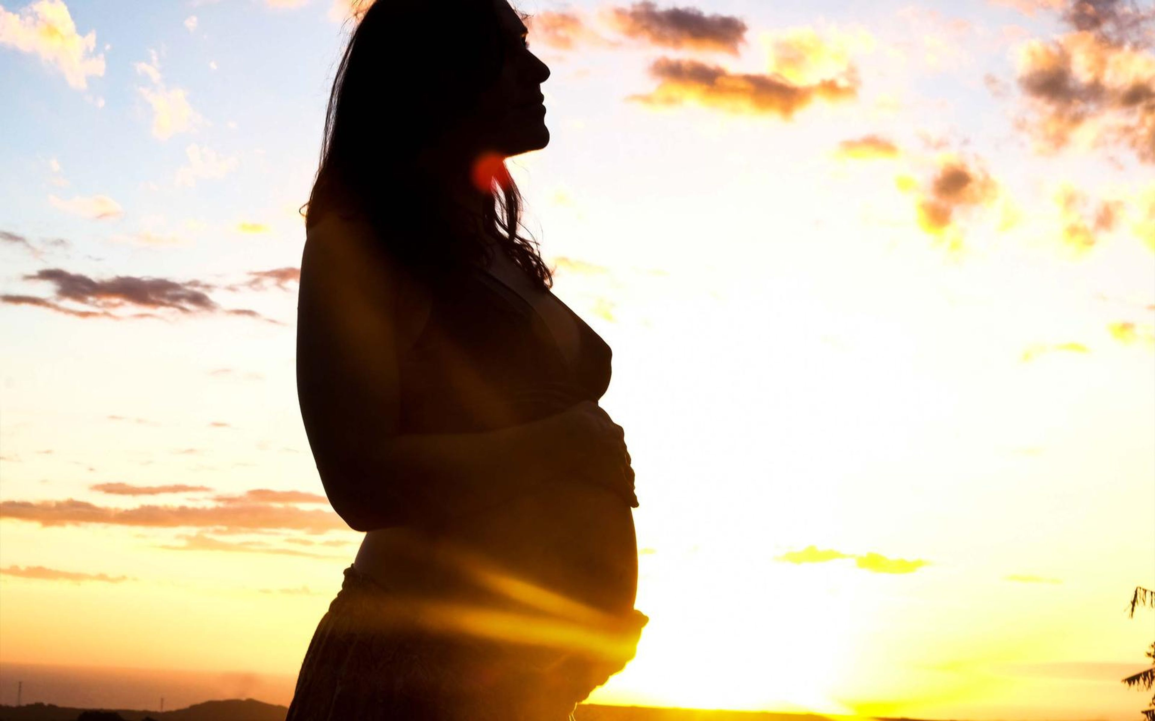 Pregnant woman in silhouette looking up, hand on her belly, bright yellow sun glow and lens flare