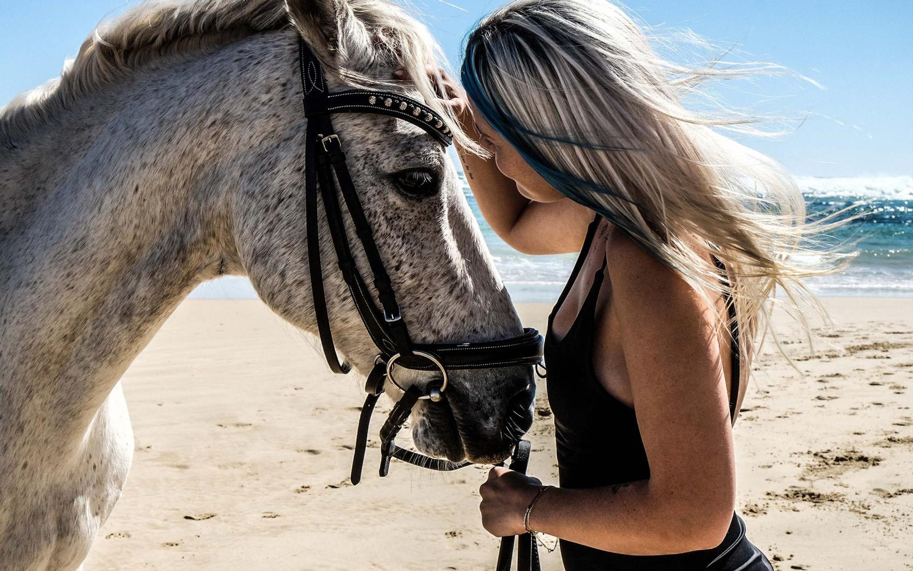Woman standing close to a white horse's head with wind lifting her hair, sea and sky behind