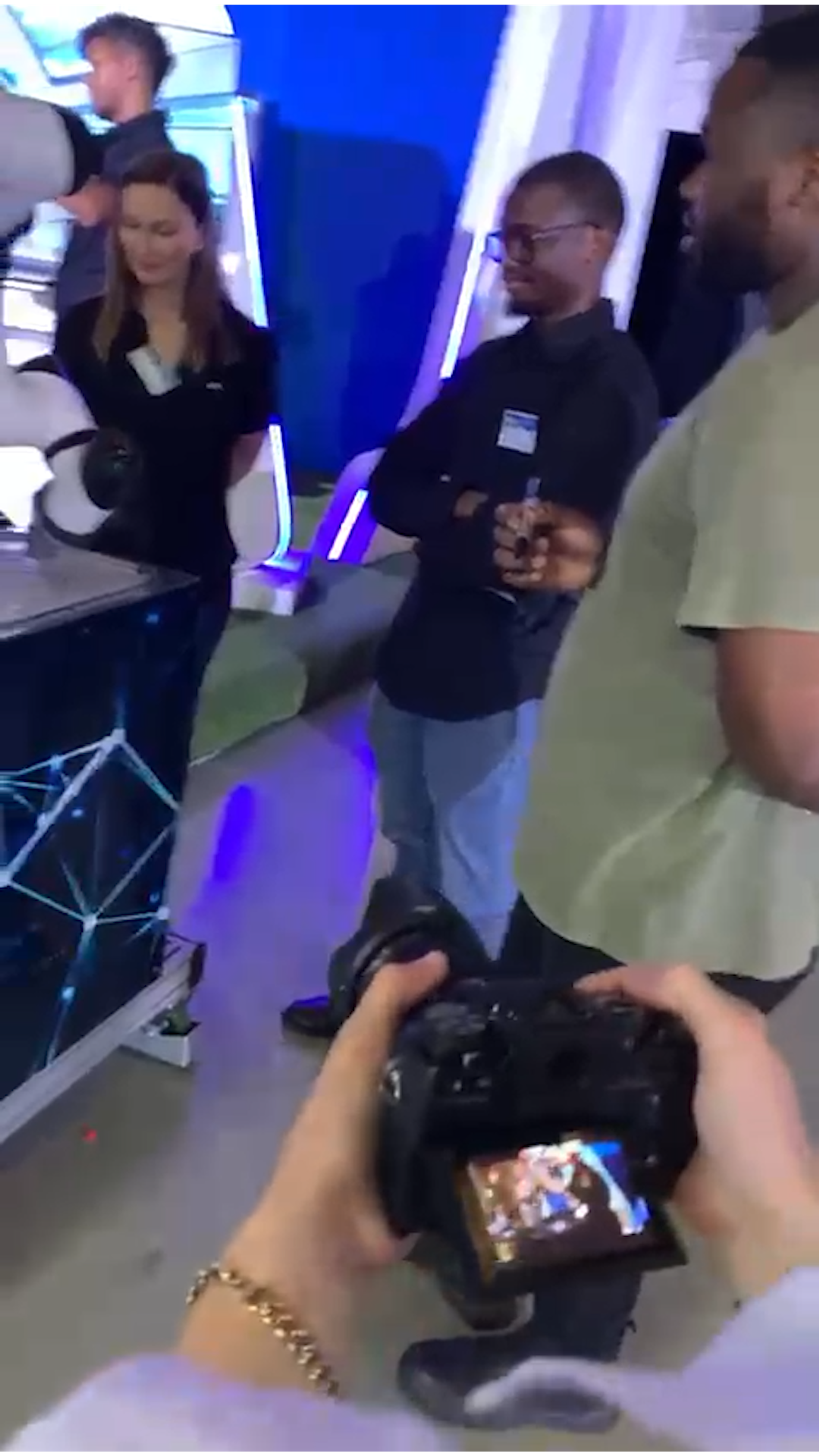 Behind the scenes at the expo, a camera screen in the foreground with visitors talking at a stand