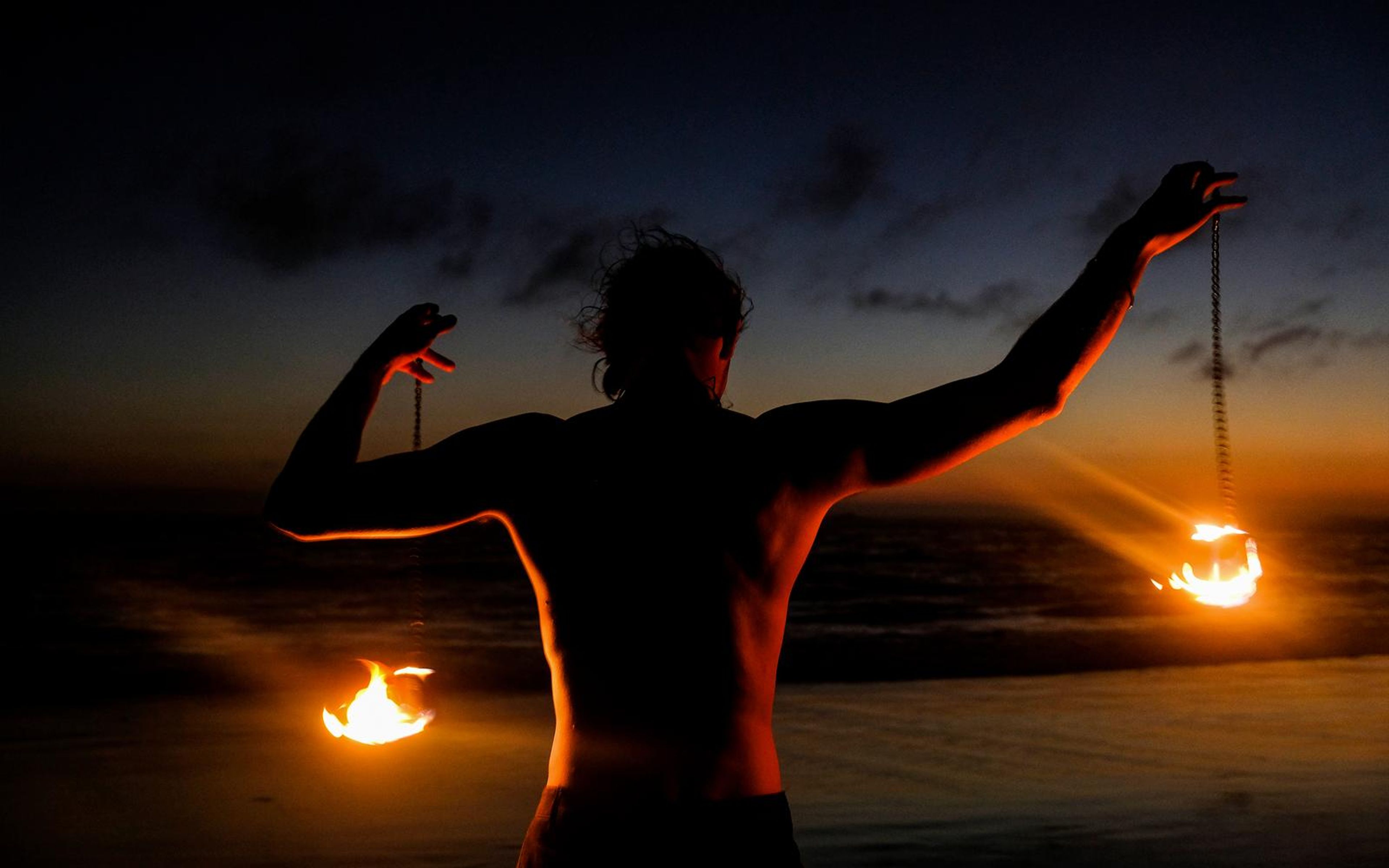 Performer from behind with arms spread and fire poi near the sand, glowing sunset above the sea