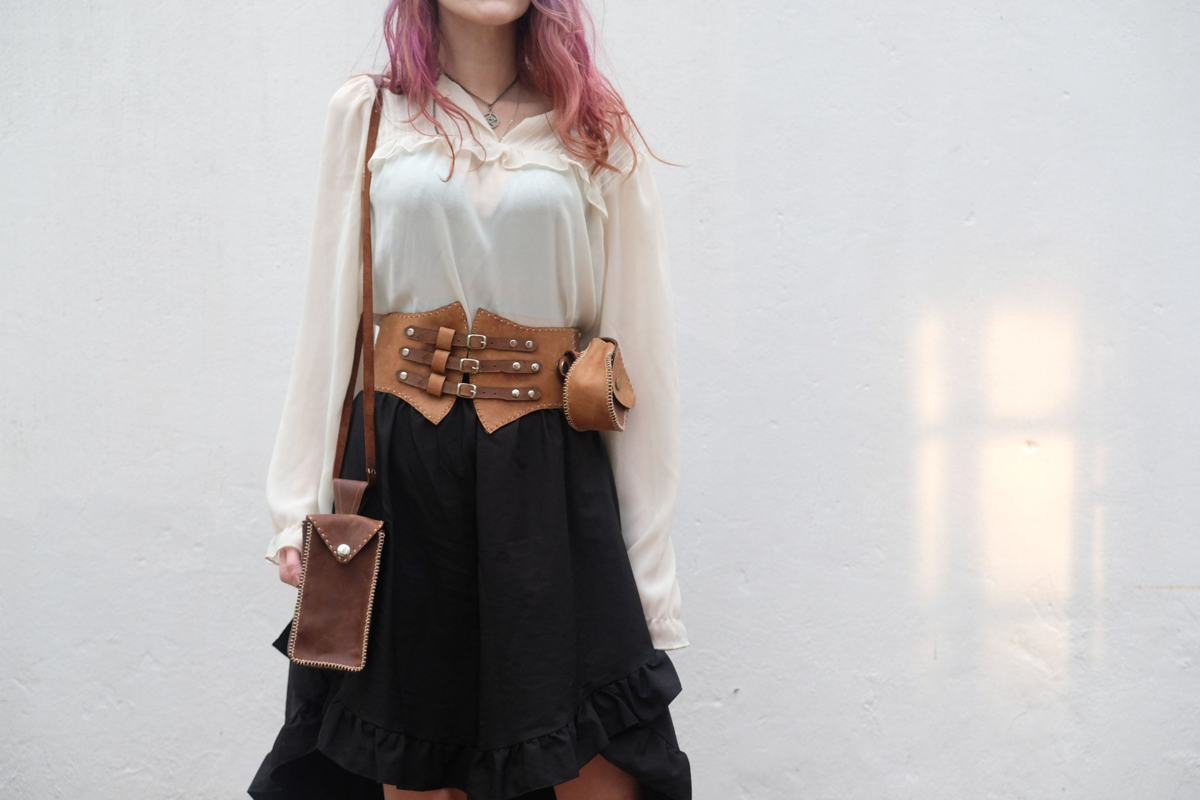 Woman standing with a leather corset belt, round pouch and a brown phone bag against a white wall