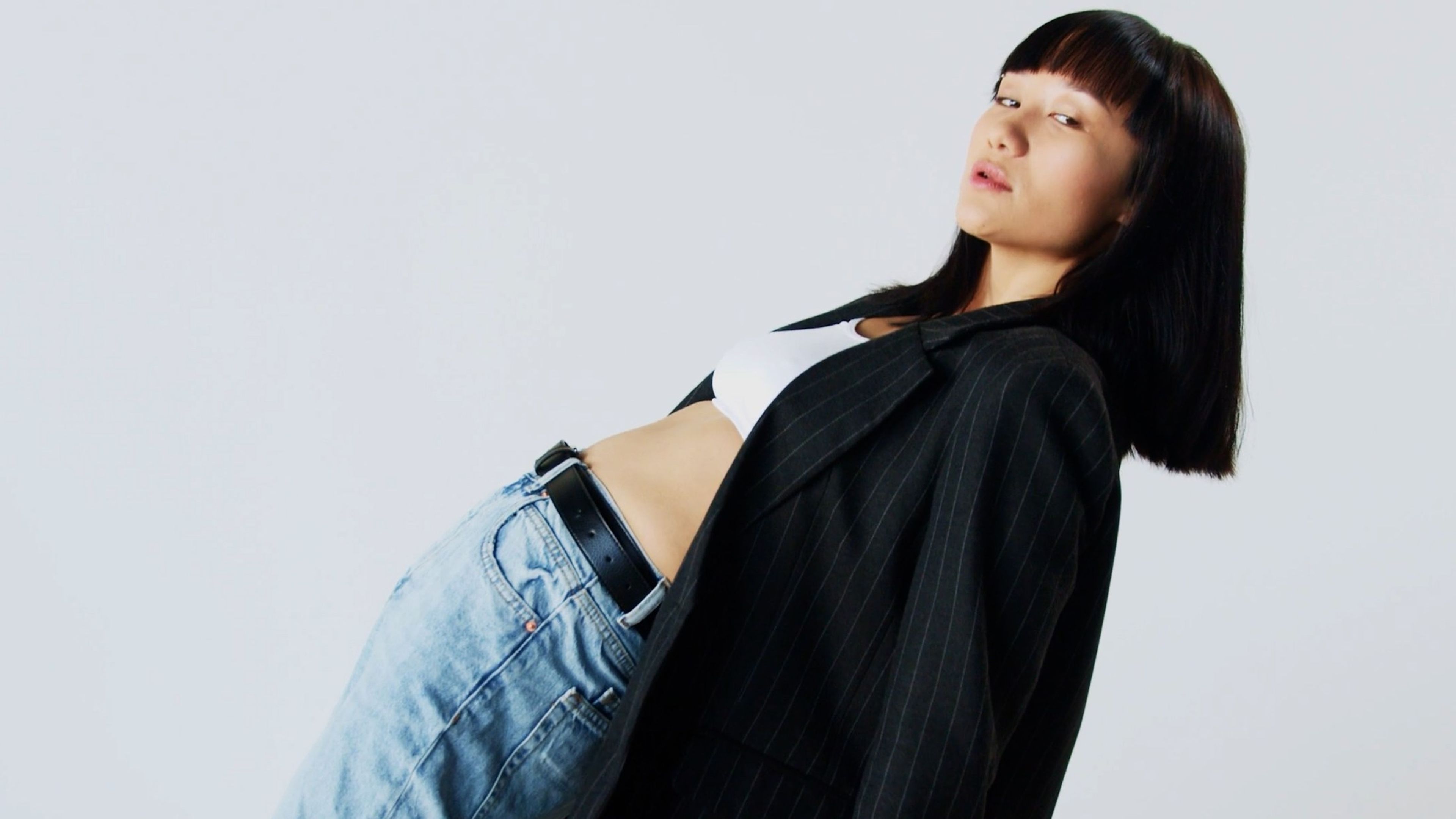 Woman in a black jacket and jeans leaning against a pale backdrop