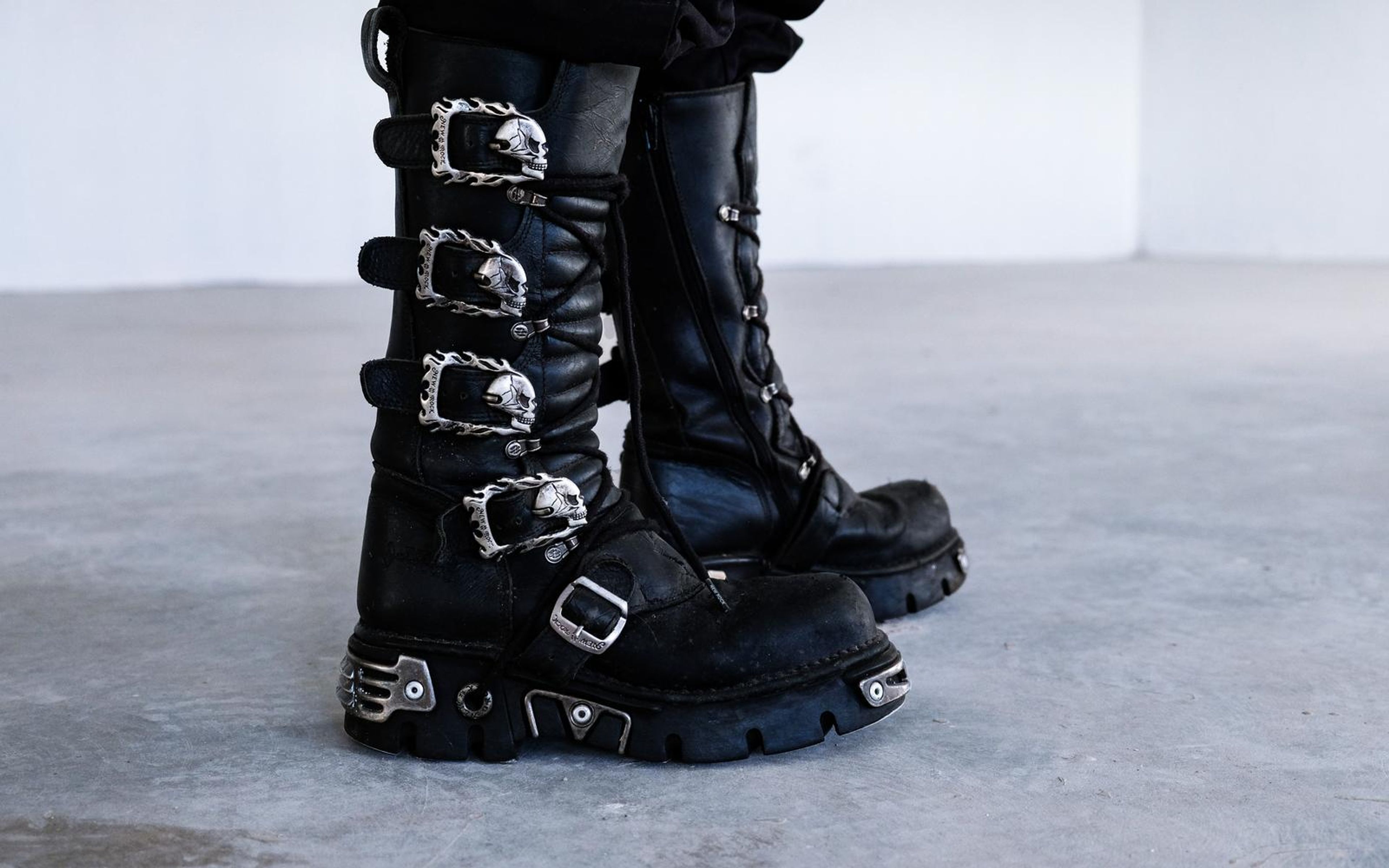 Pair of black boots with skull-shaped buckles standing on a concrete floor against a white wall