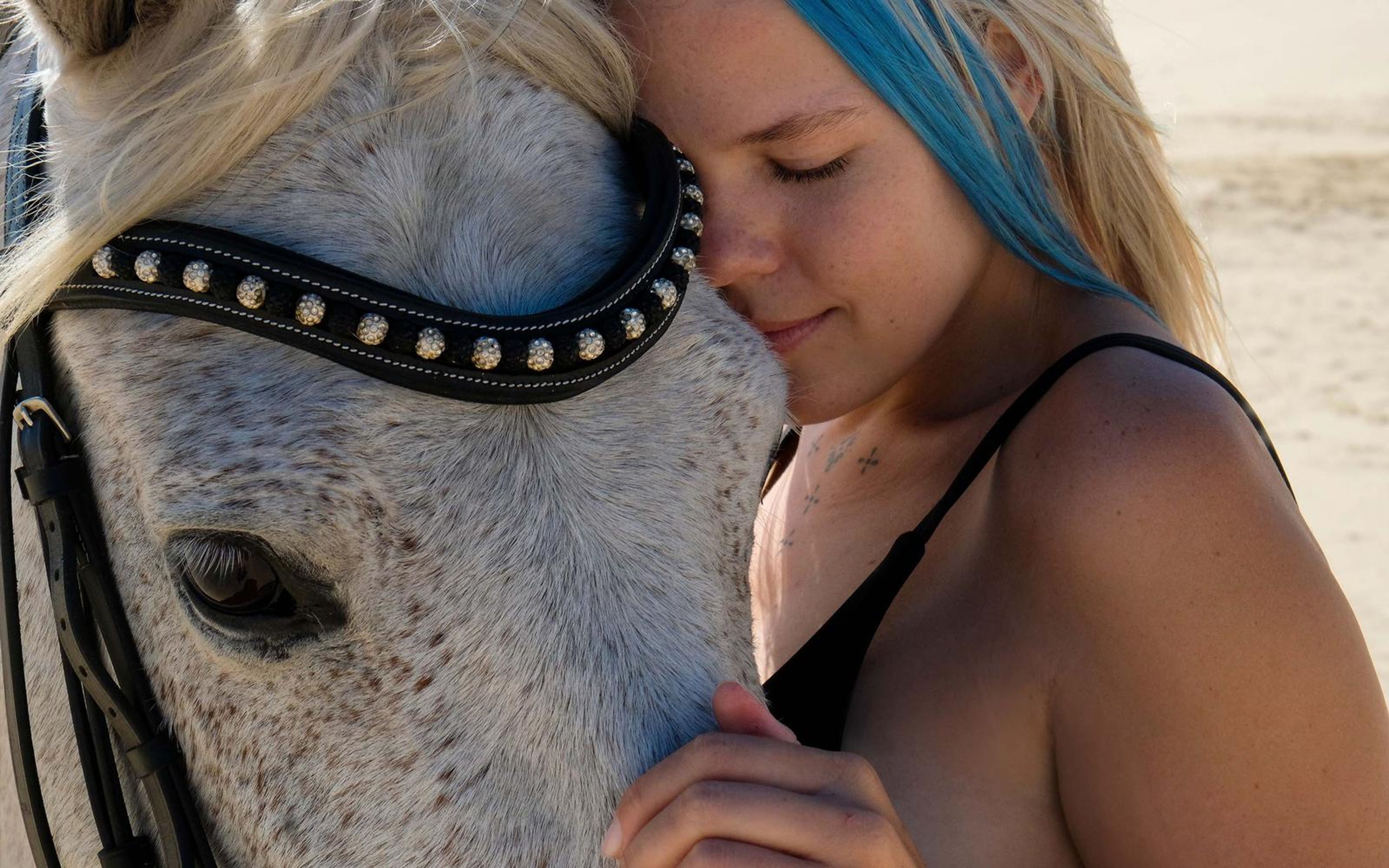 Woman with blue-streaked hair pressing her forehead to a white horse's face, eyes closed