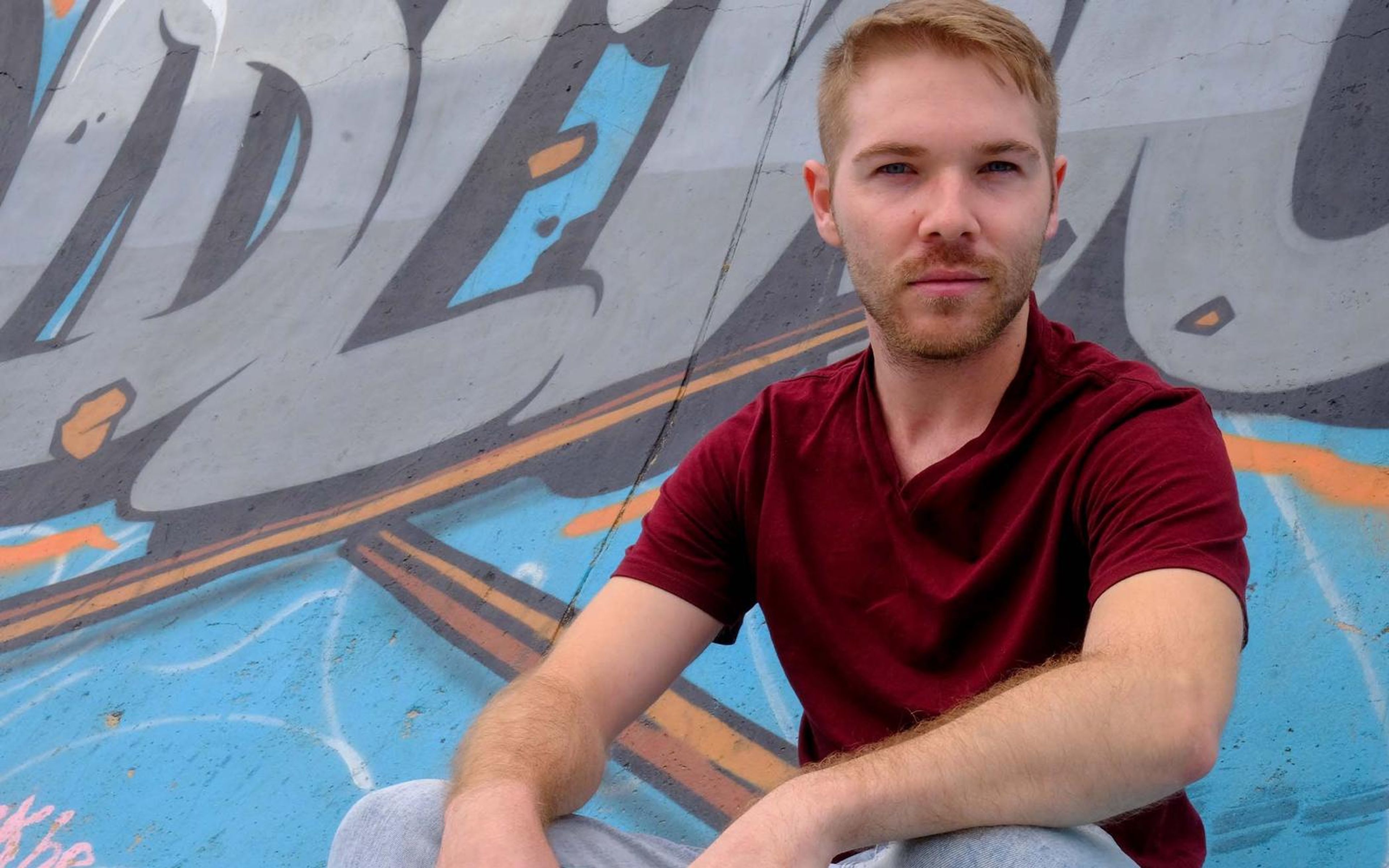 Man with a direct gaze seated in front of large graffiti letters, maroon tee, blue painted floor