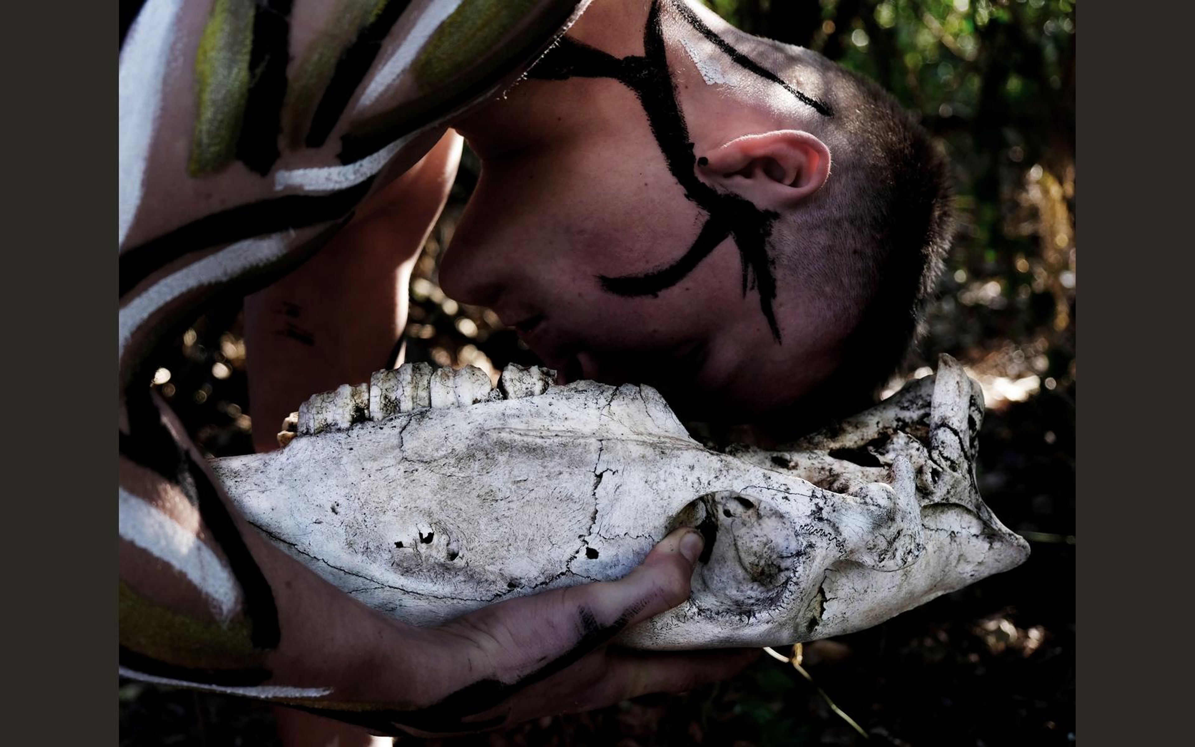 Performer kissing the jaw of a bleached bull skull, with black and white face paint and forest greens behind