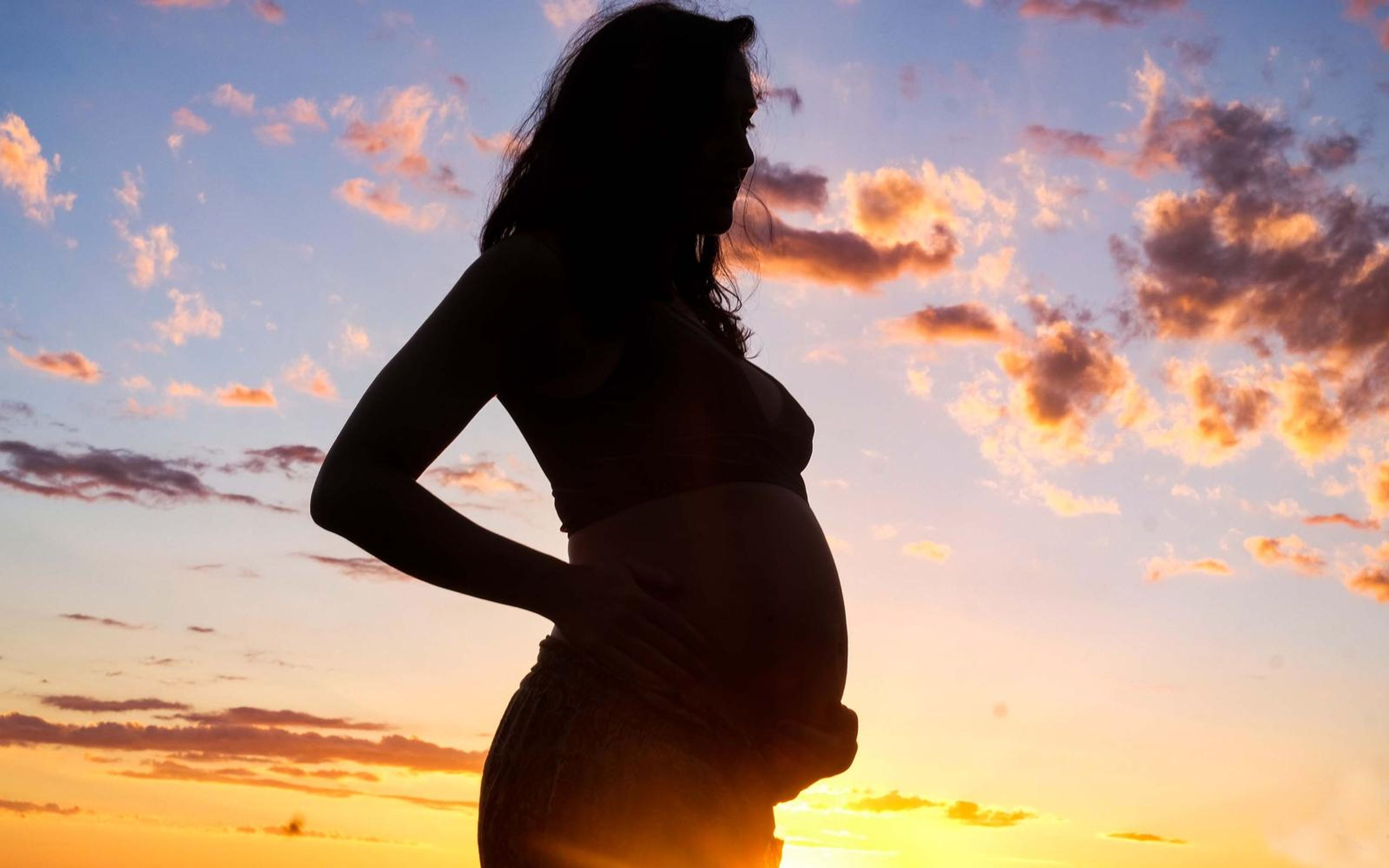 Silhouette of a pregnant woman, hand on her lower back, lilac and orange sunset sky
