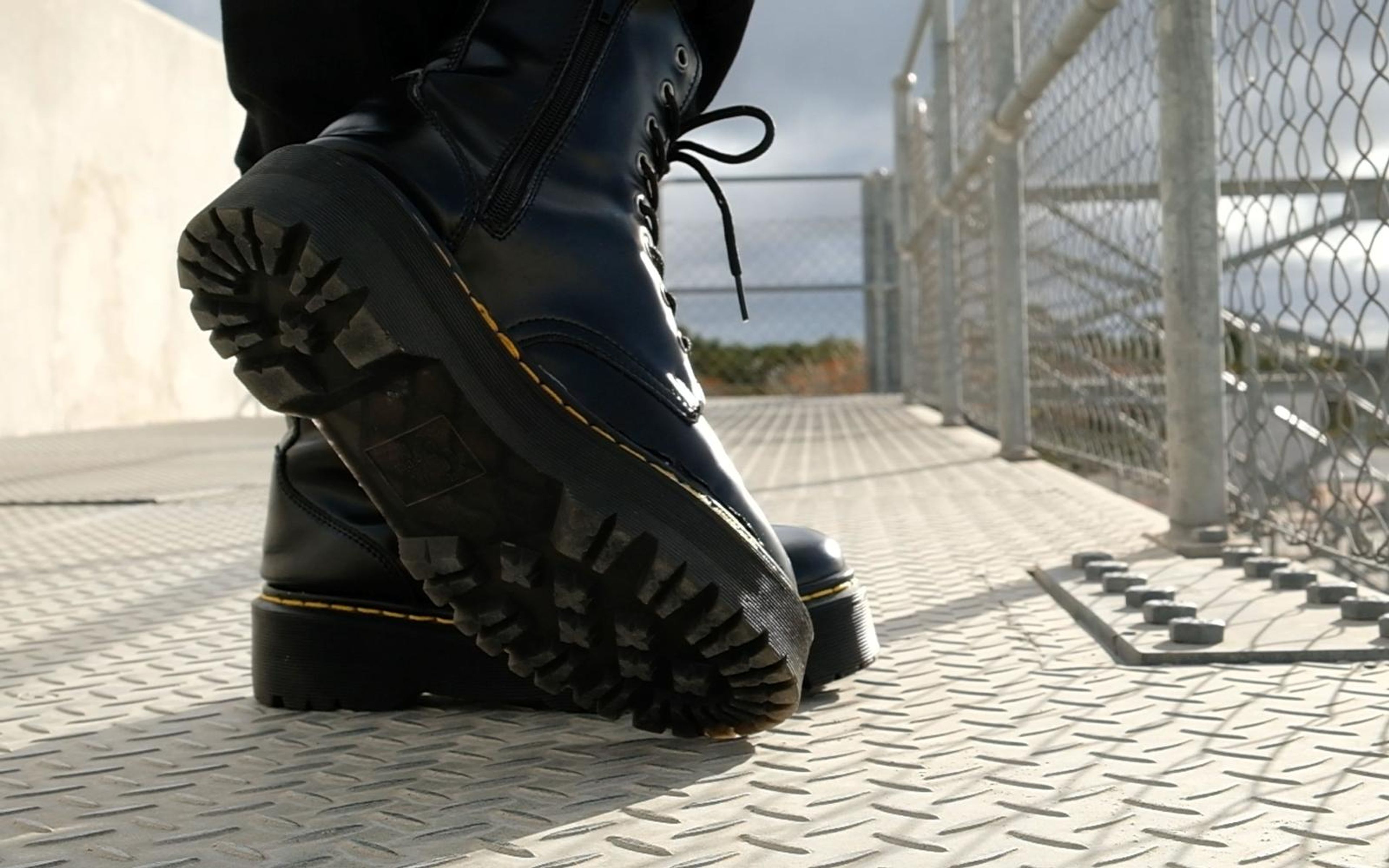 One black boot lifted to show its thick treaded sole, other boot planted, metal mesh and sky behind