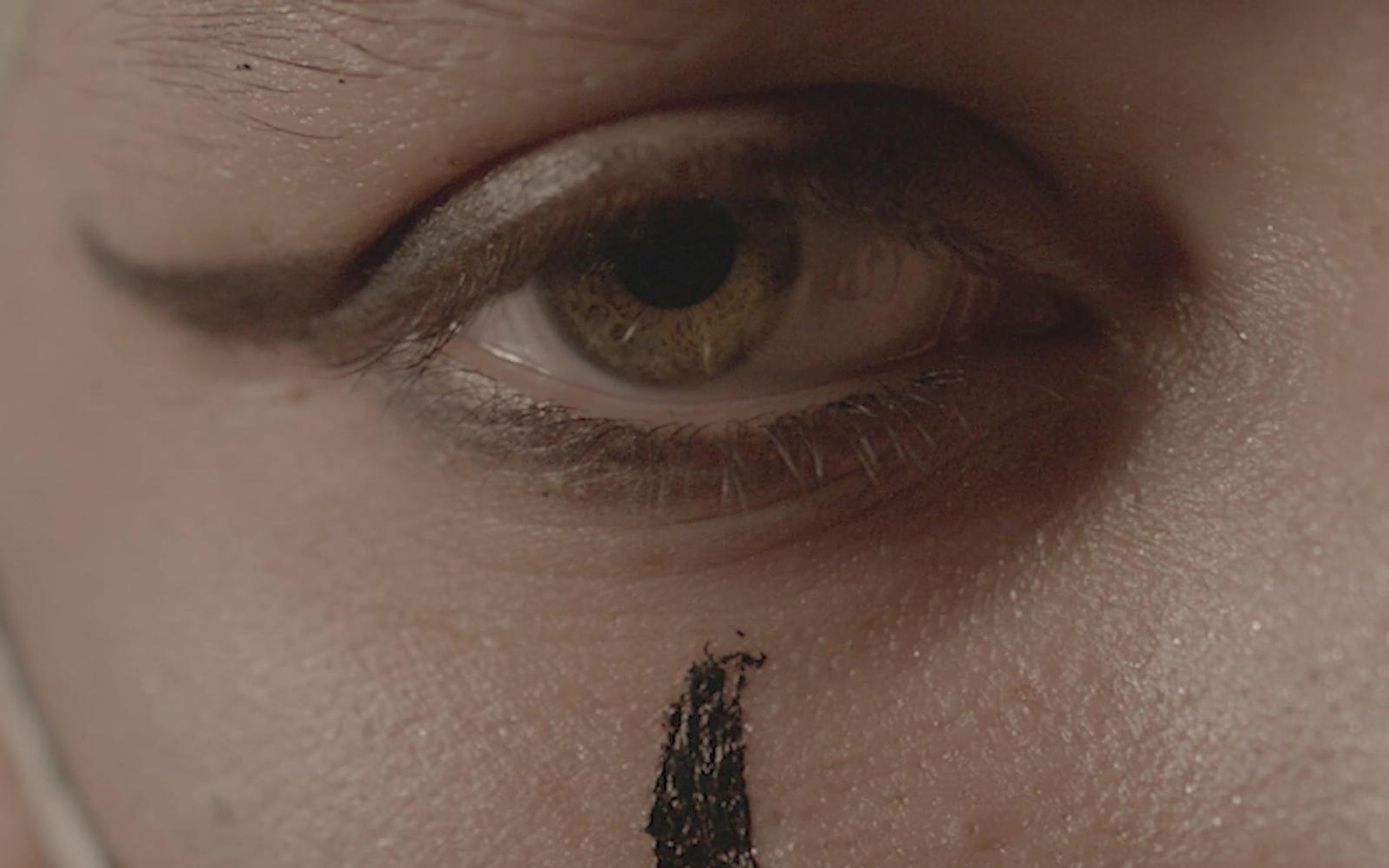 Extreme close-up of an eye with winged liner and a black paint mark below it, in muted skin tones