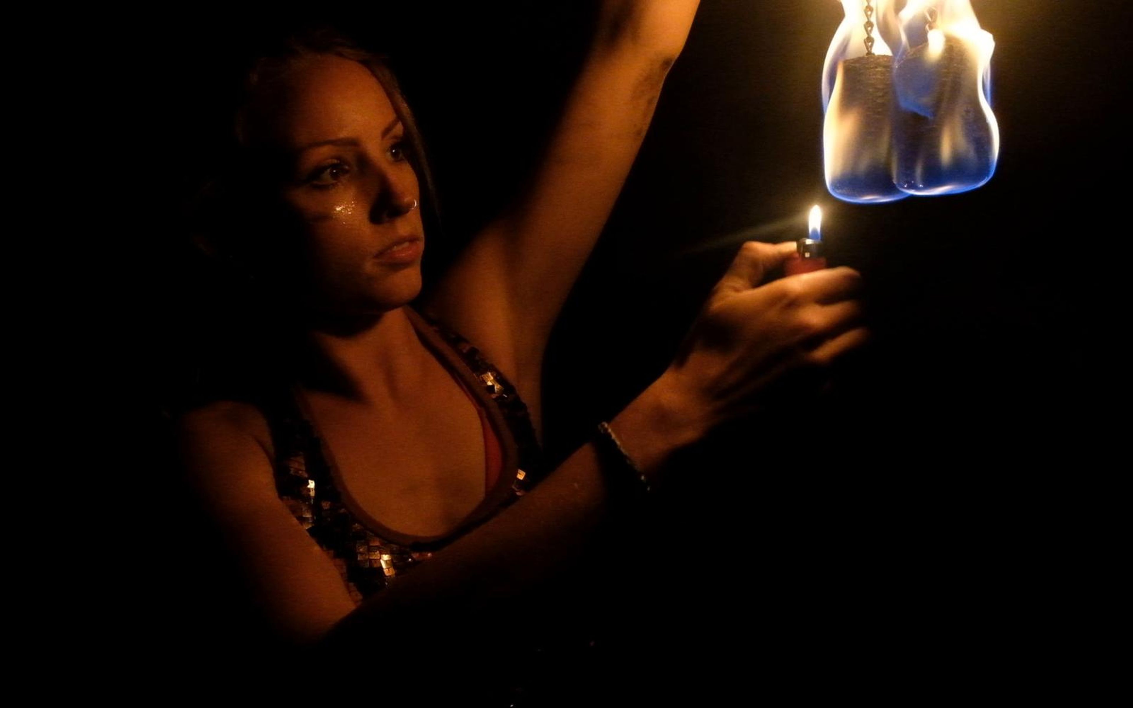 Woman in a sequinned top lighting a fire prop with a lighter against a dark background