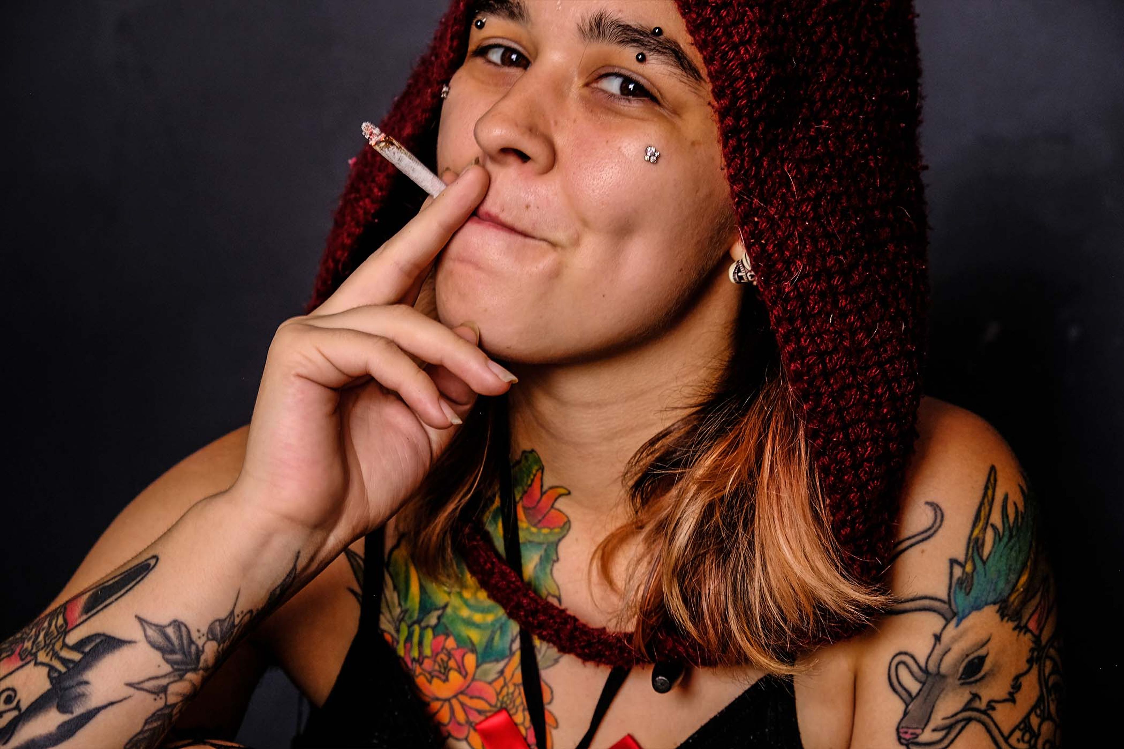 Close-up portrait, subject in a maroon knit hood smoking a cigarette with a faint smile, tattoos visible on the shoulder