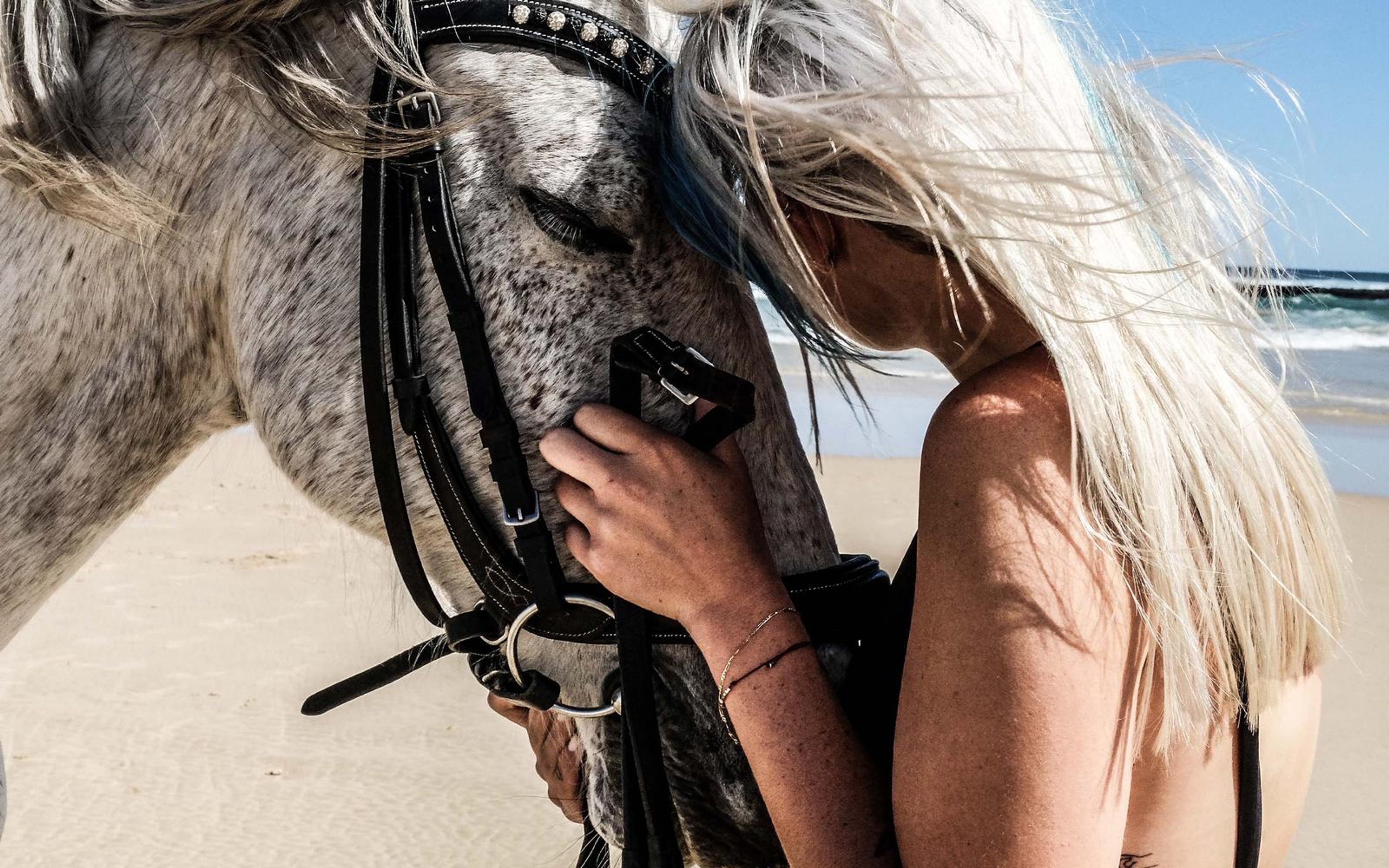 Woman in a swimsuit with a hand on a horse's bridle and her hair blowing across the horse's face