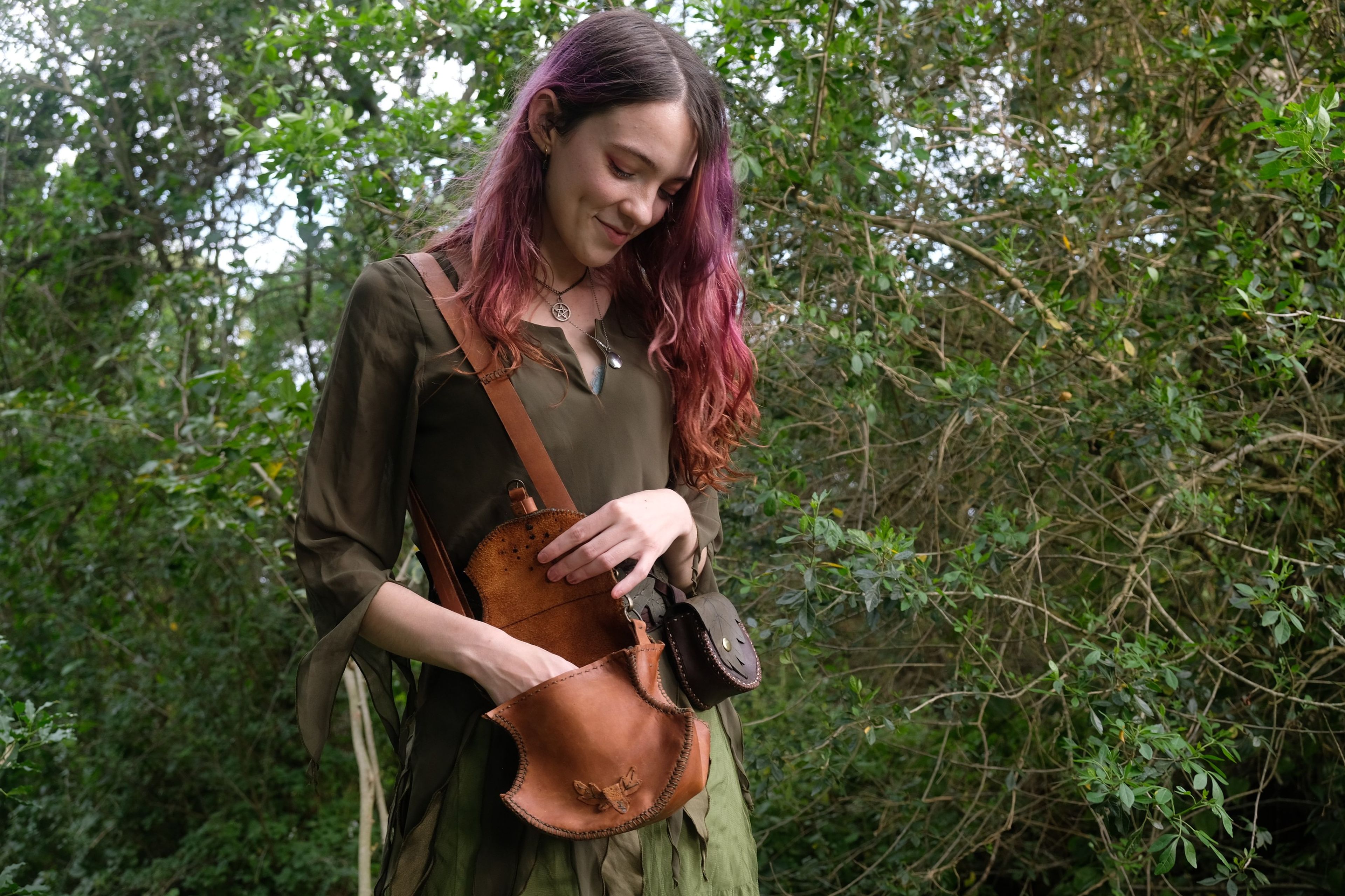 Woman smiling down at a tan leather bag, standing among green shrubs