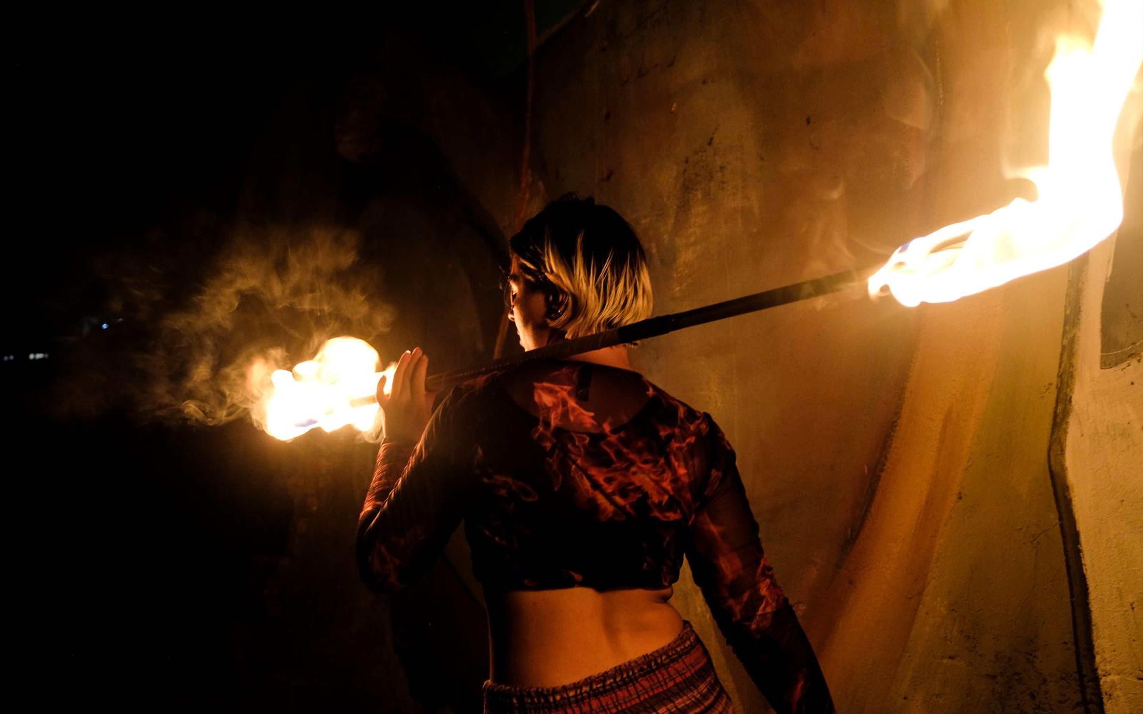 Performer seen from behind with a fire staff across the shoulders, a flame at each end and smoke at left