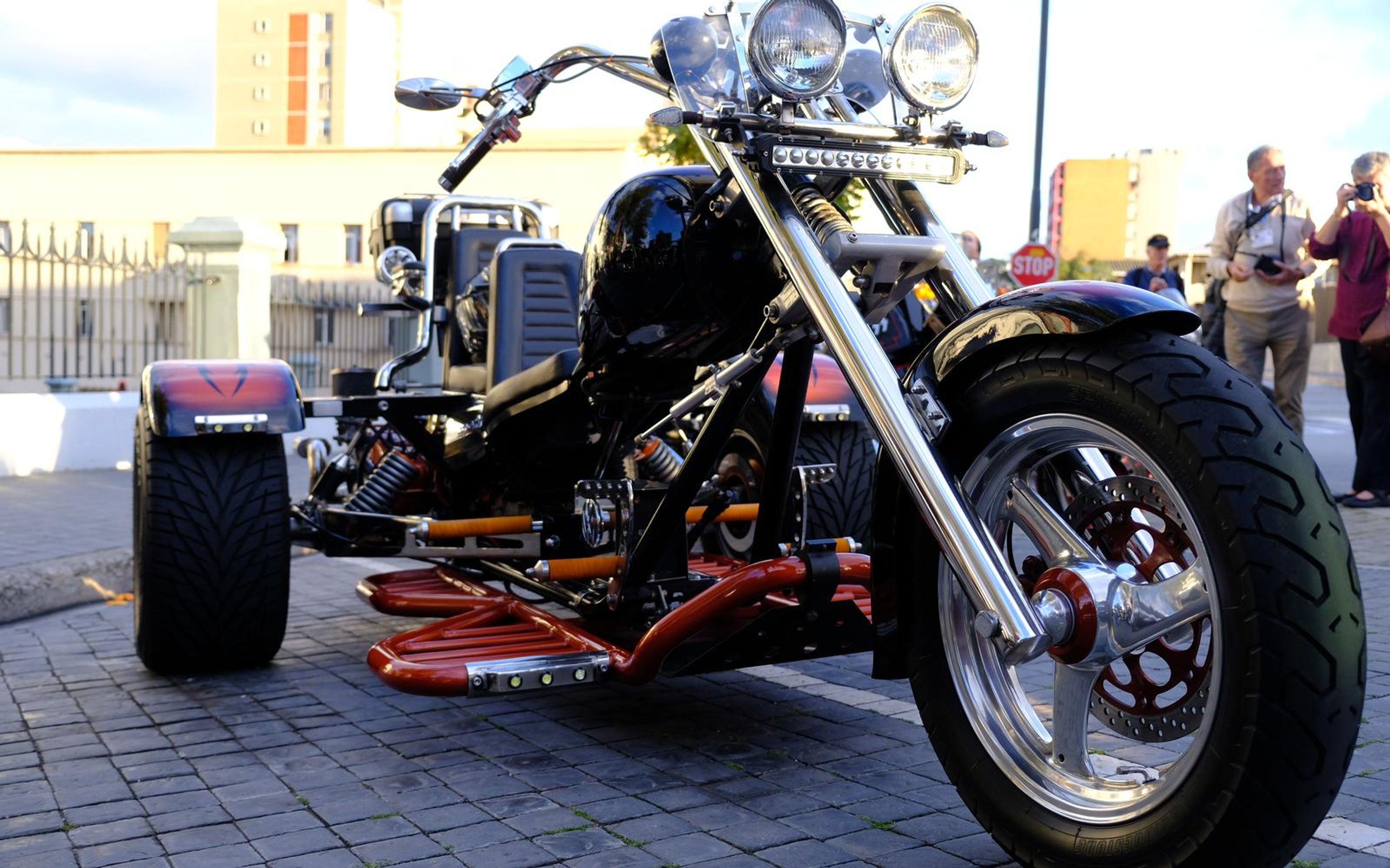 Low front three-quarter view of a black chopper-style trike with chrome forks, red footboards and a wide rear tyre on cobblestones