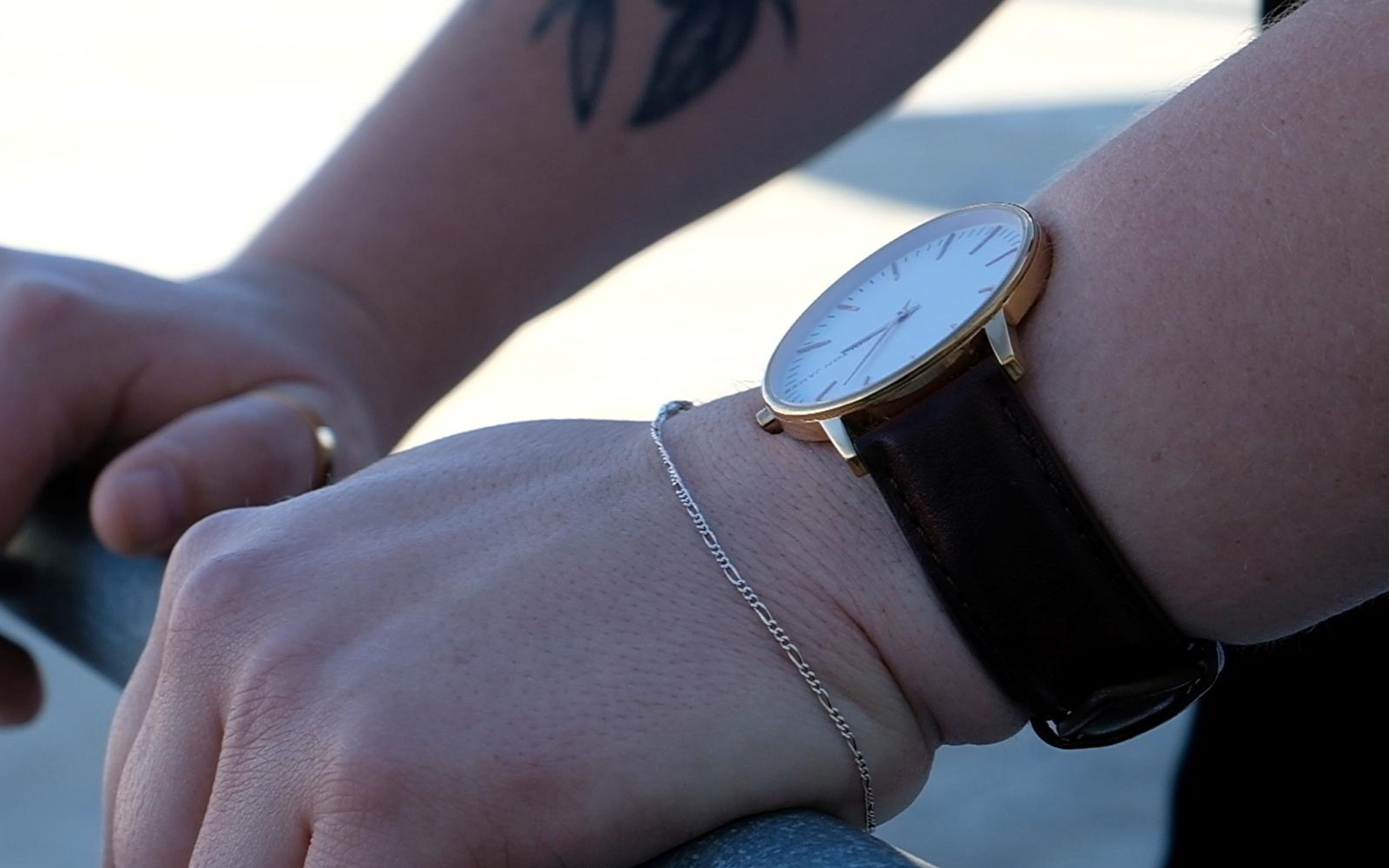 Watch and silver bracelet on a wrist, hand gripping a metal rail, small tattoo on the forearm, bright background