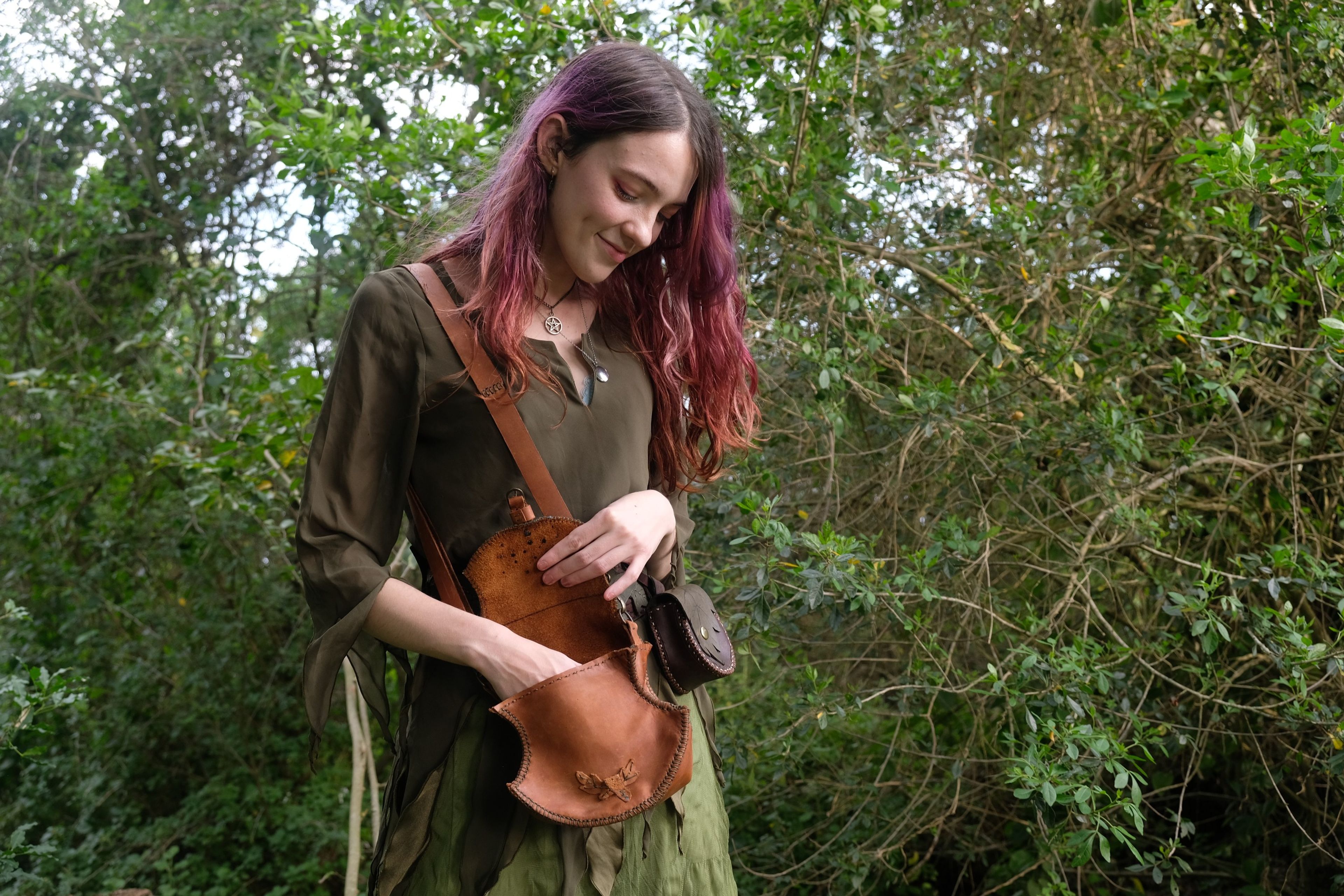 Woman in a green dress opening a tan leather bag among shrubs