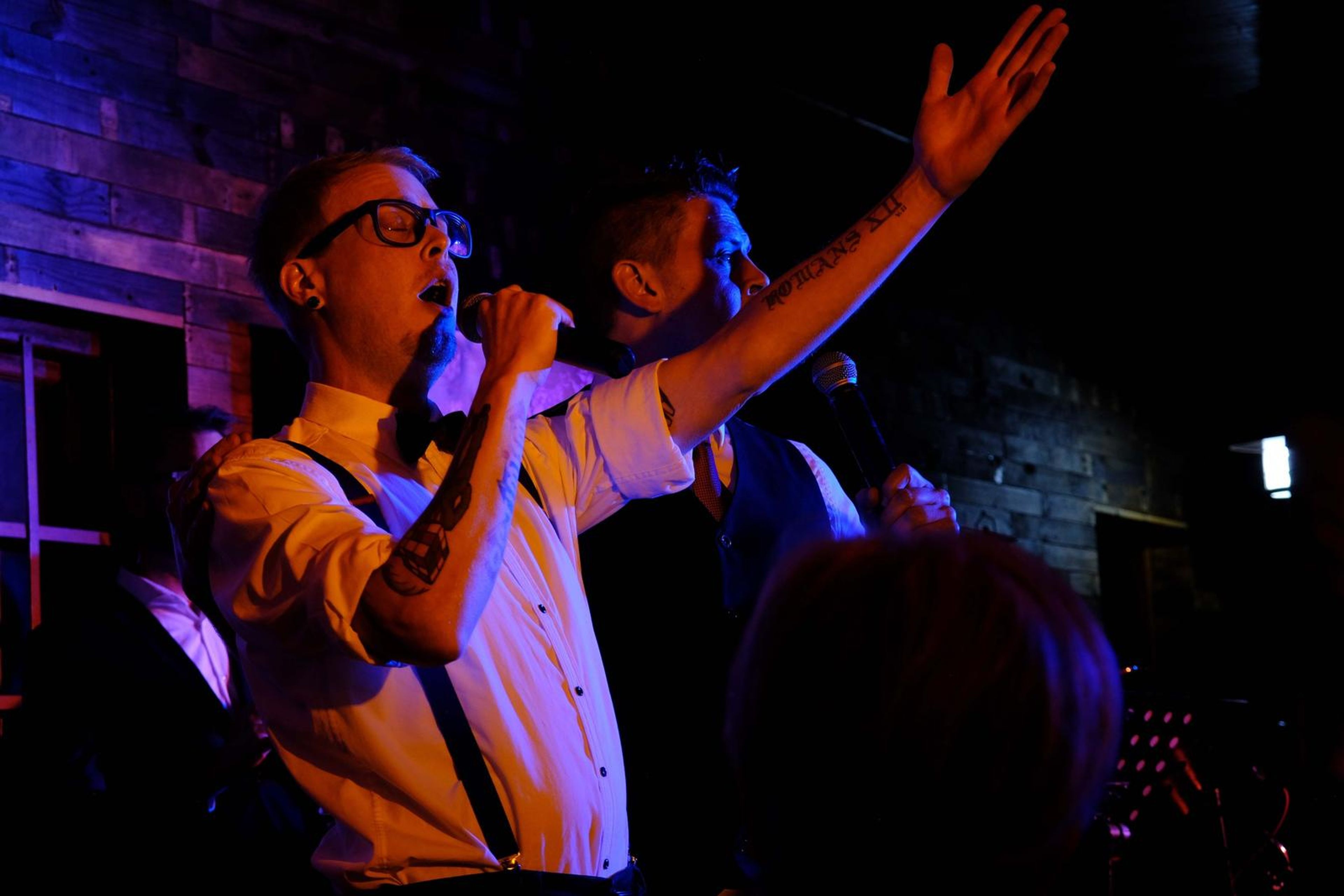 Singer in glasses and suspenders raising his arm as he sings