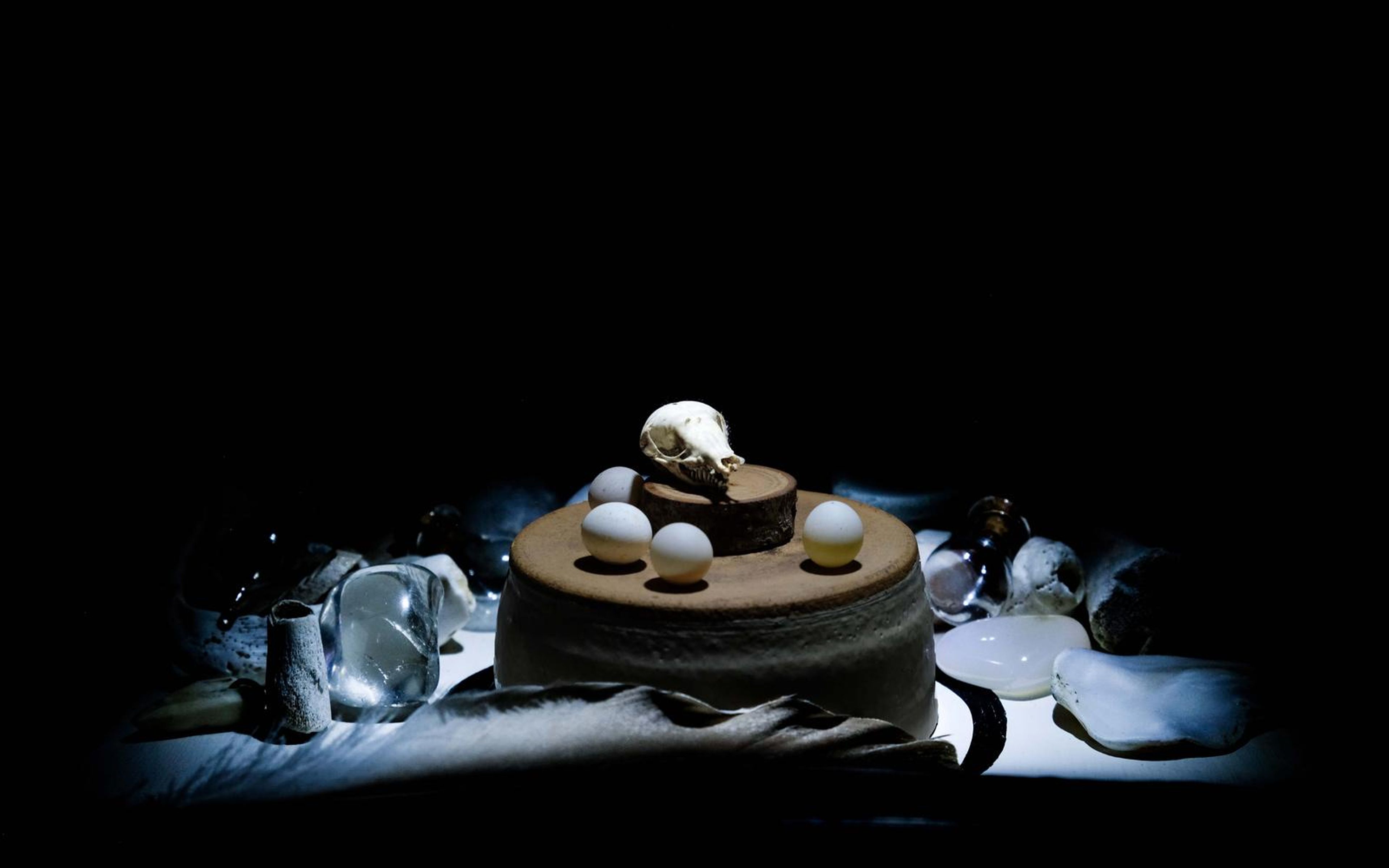 Small skull and white spheres on a round wooden stand, side-lit against darkness with glass and stones behind
