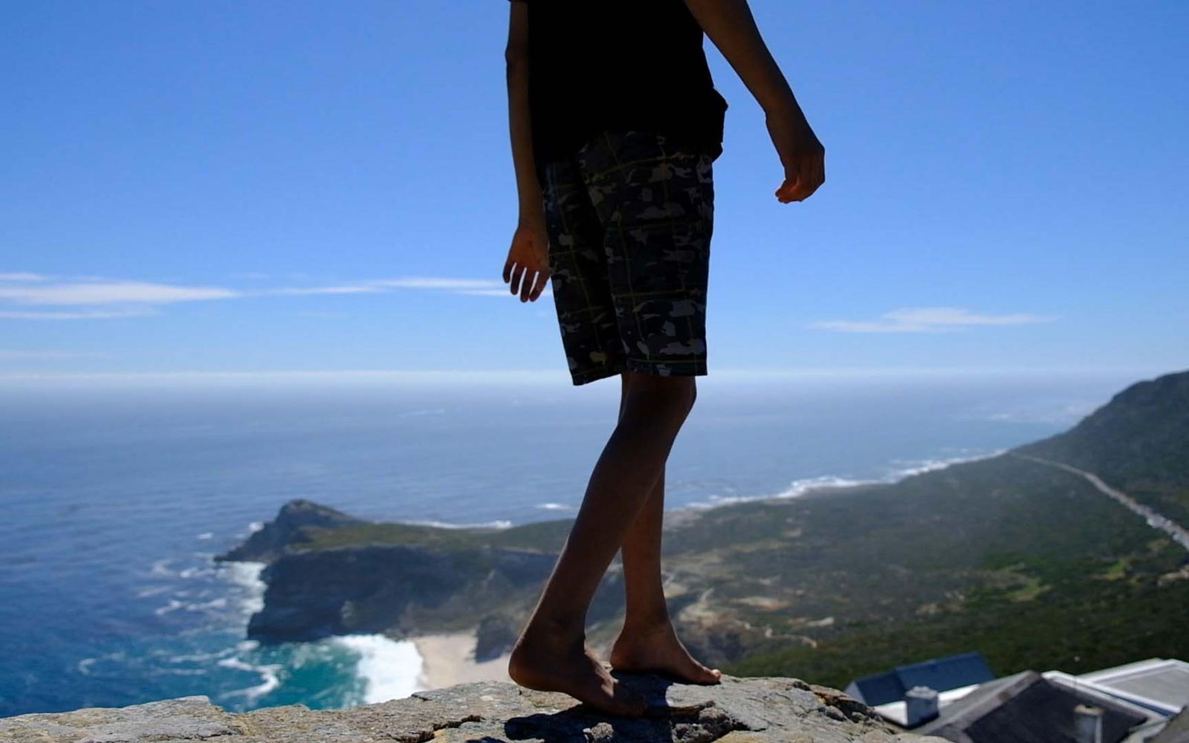 Bare feet and legs of a person stepping along a rocky ledge high above a sunlit coastline
