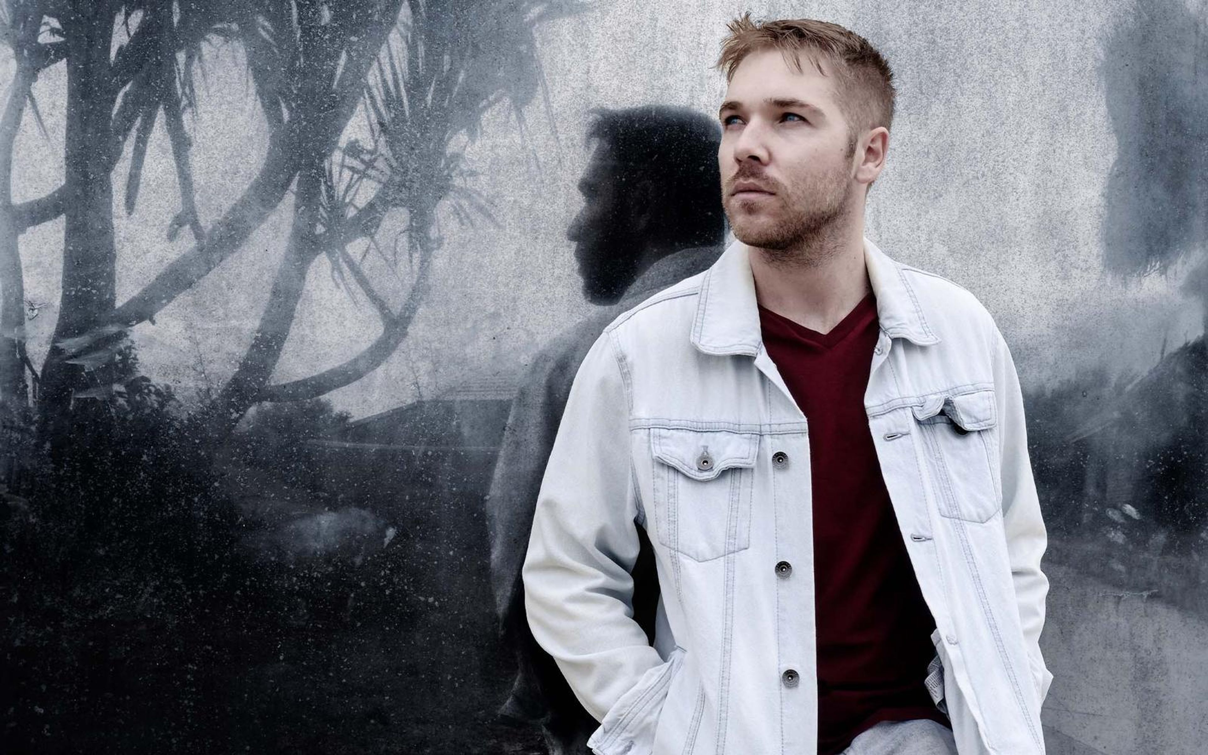 Man in a pale denim jacket looking off to the side beside a dark leaf shadow on a grey wall, desaturated