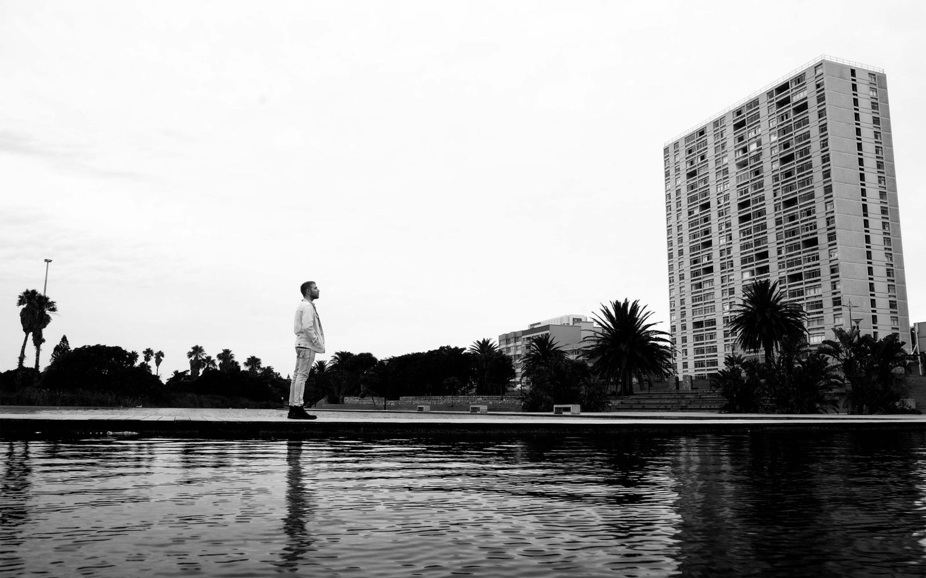 Black and white wide shot of a man standing at a pool edge with palm trees and a tall apartment block behind