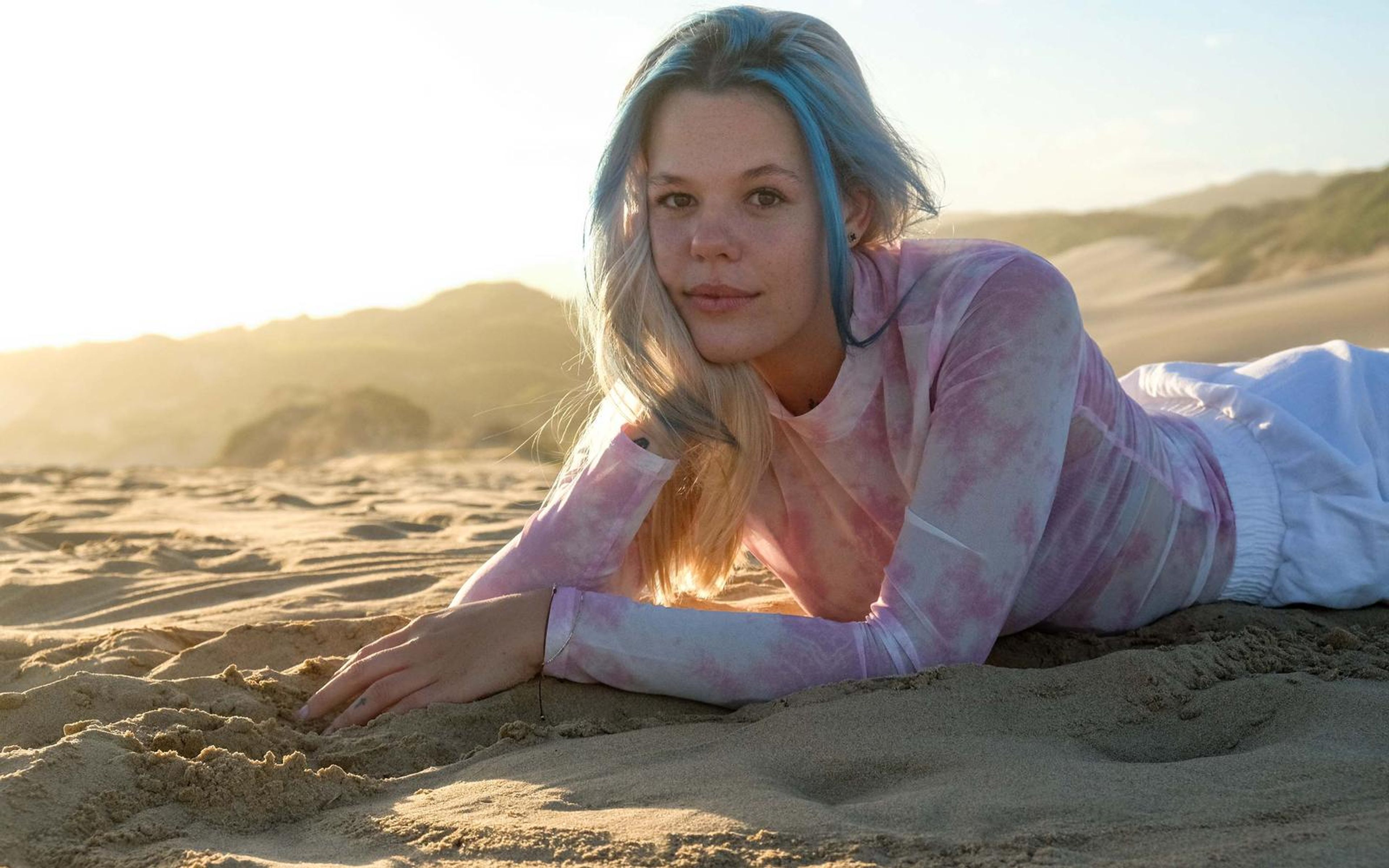 Woman with blue-streaked hair lying on the sand propped on her arms, smiling at the camera, dunes behind