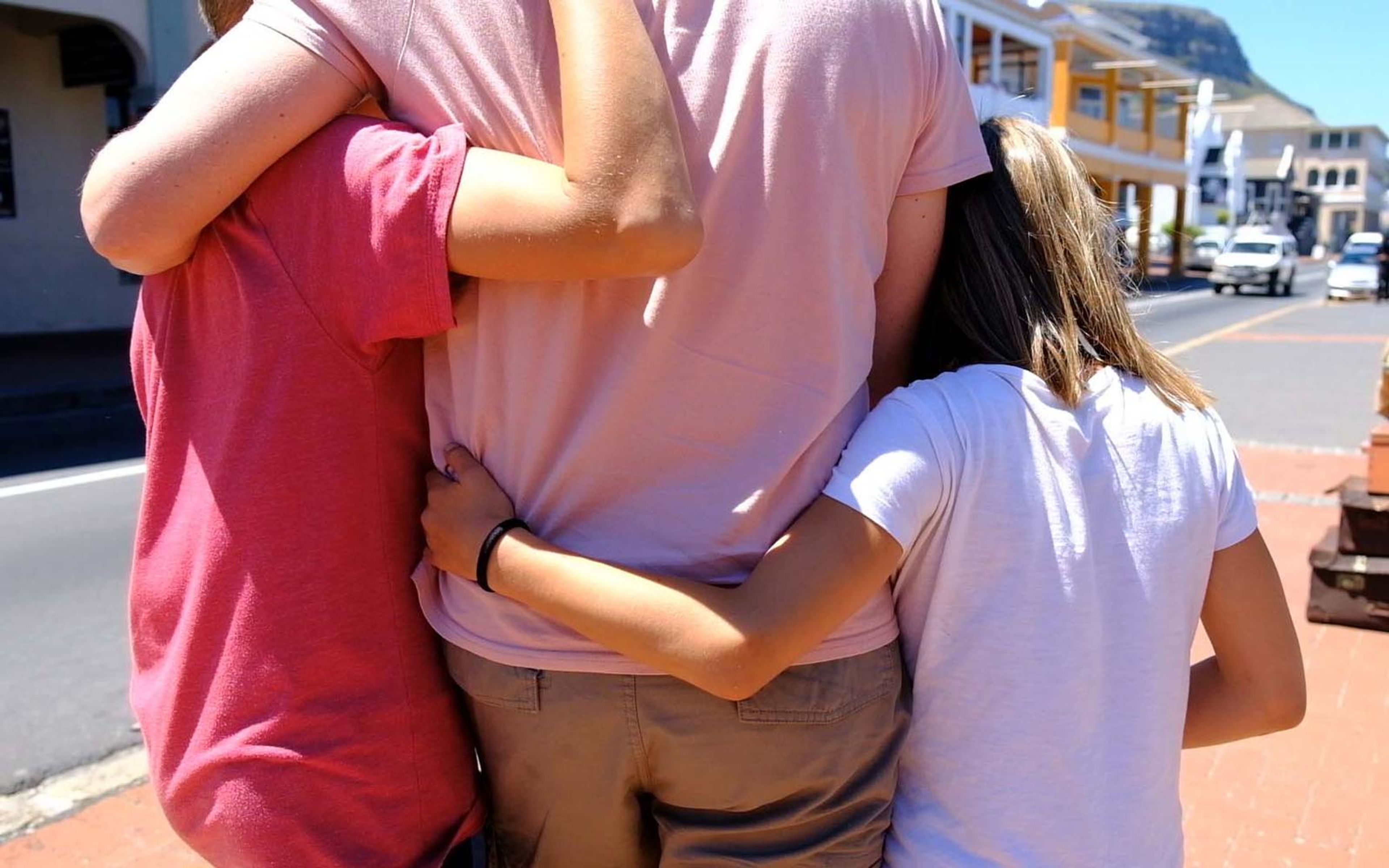 Two children and an adult hugging with their backs to the camera on a sunny street with shopfronts