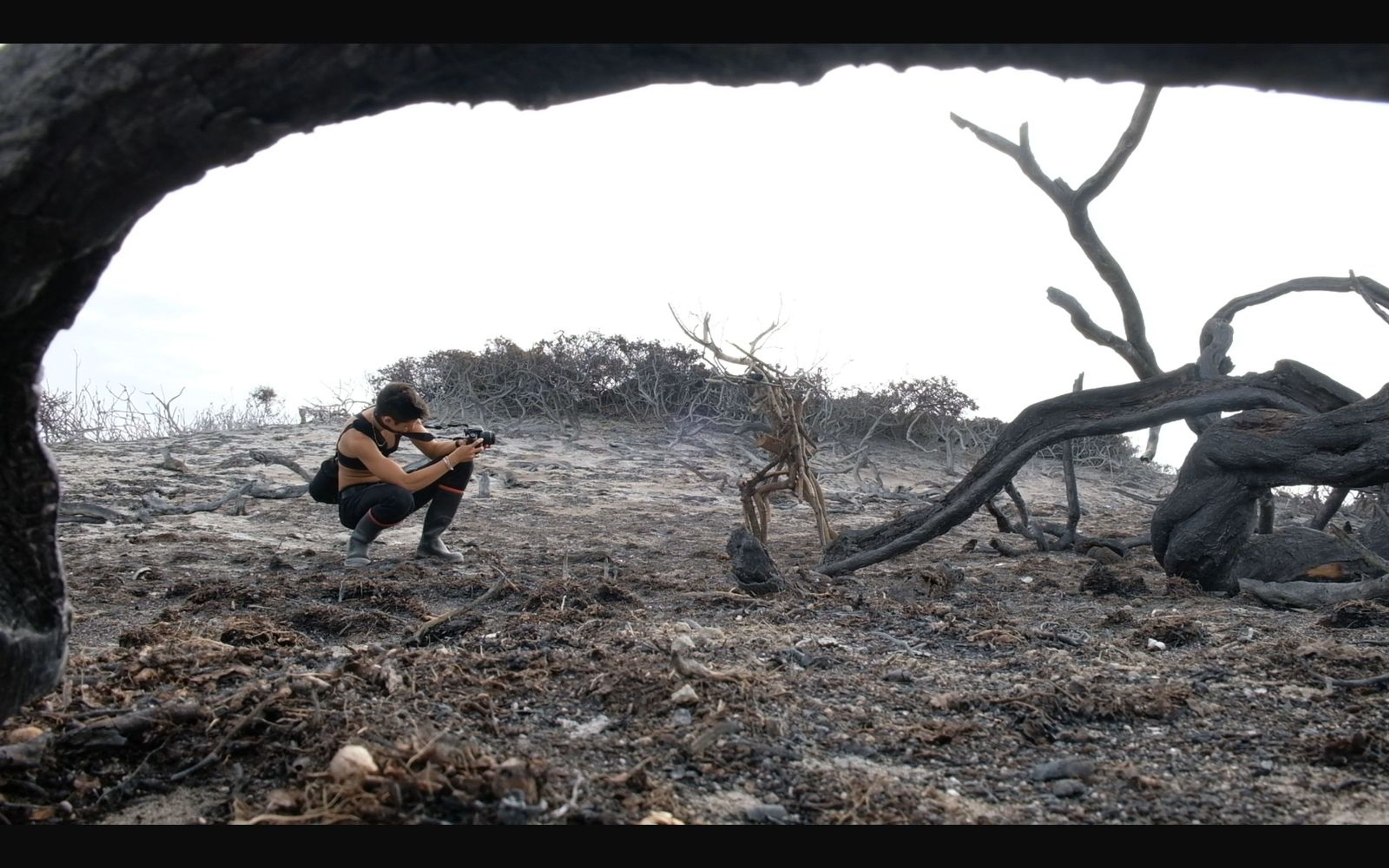 Small crouched figure photographing the ground through a dark burnt-wood arch onto grey ash
