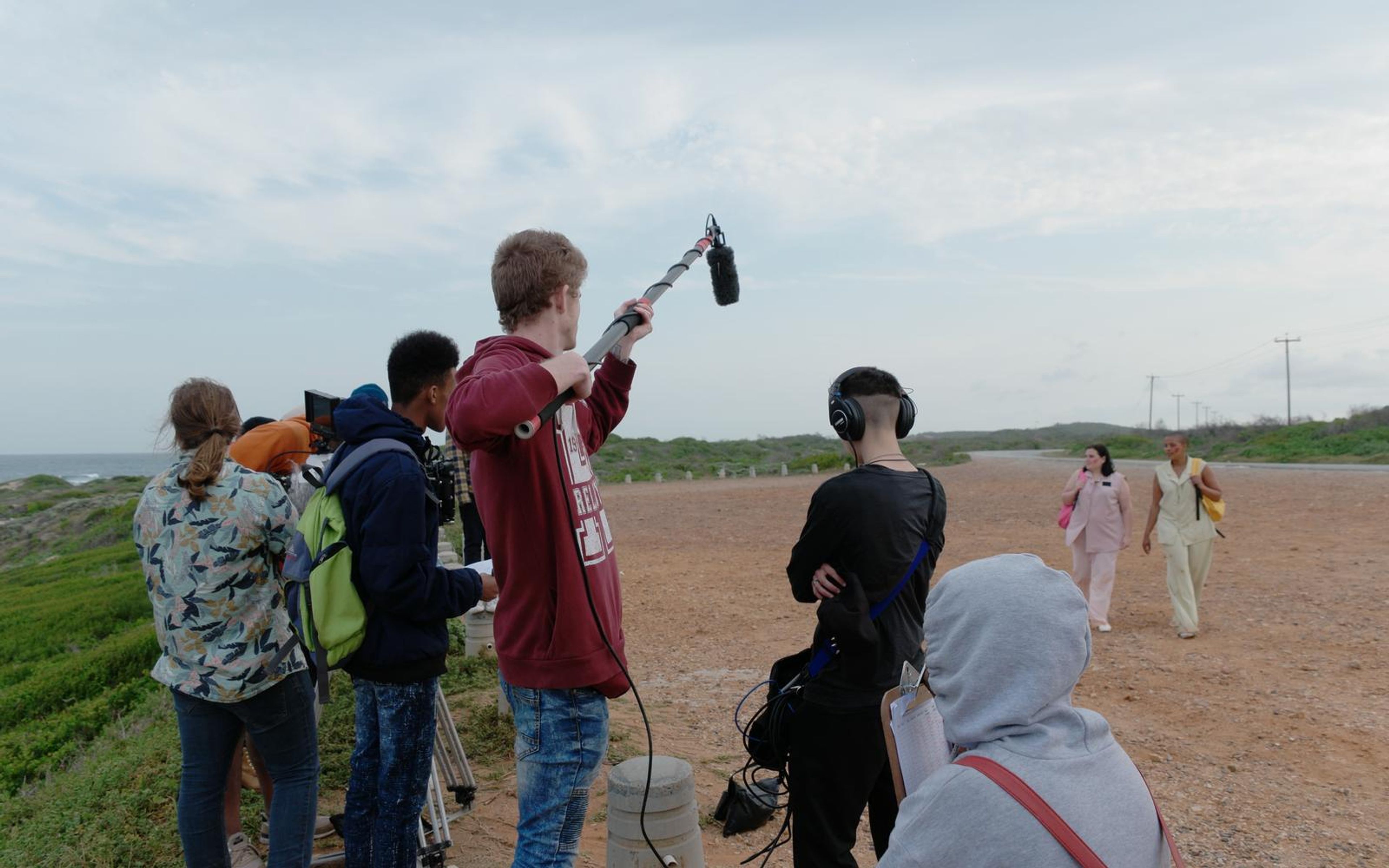 Boom operator in a maroon hoodie raising a pole on a coastal gravel lot with crew standing behind