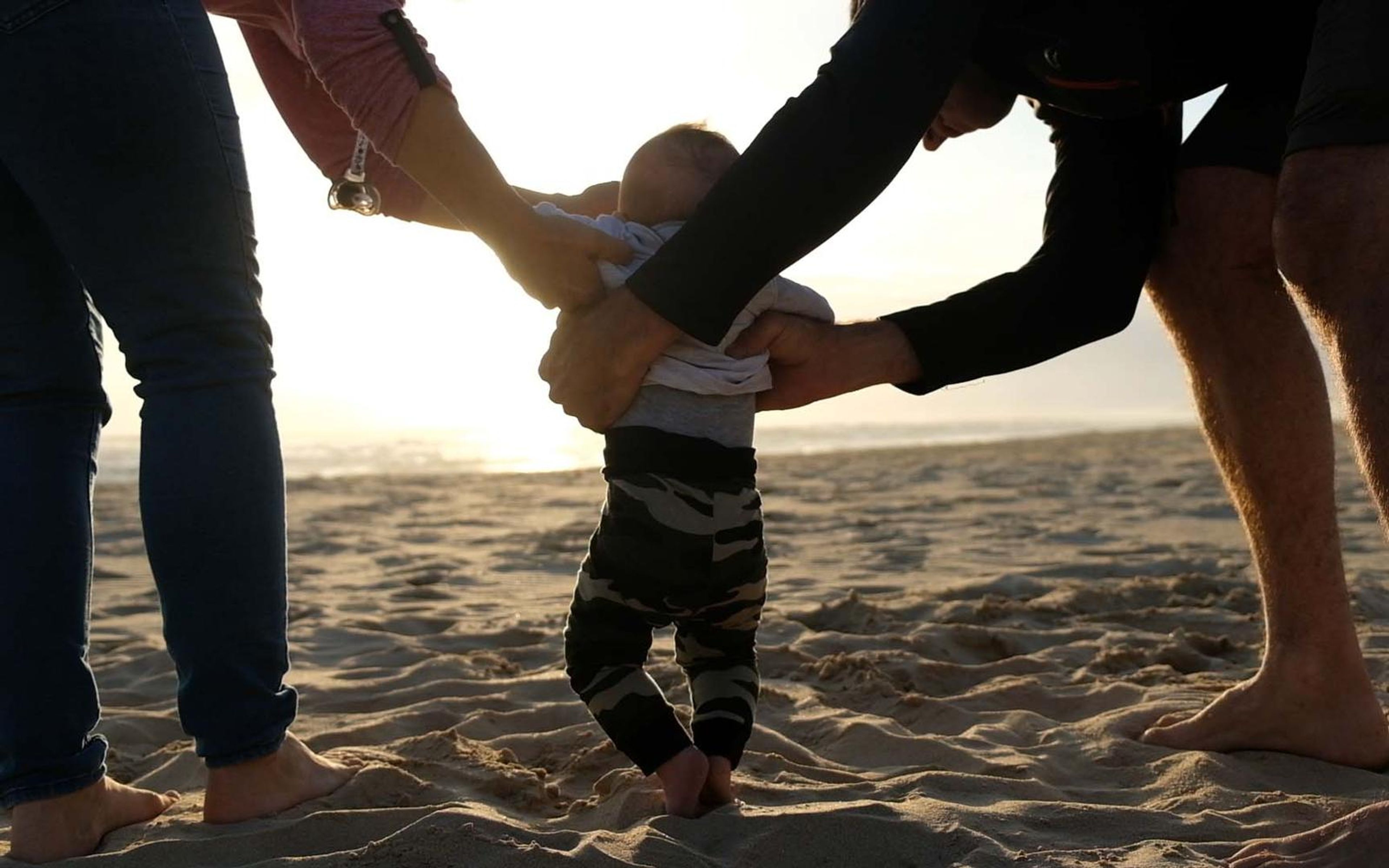 Two adults steadying a toddler in camouflage trousers as he walks across sand in low sun