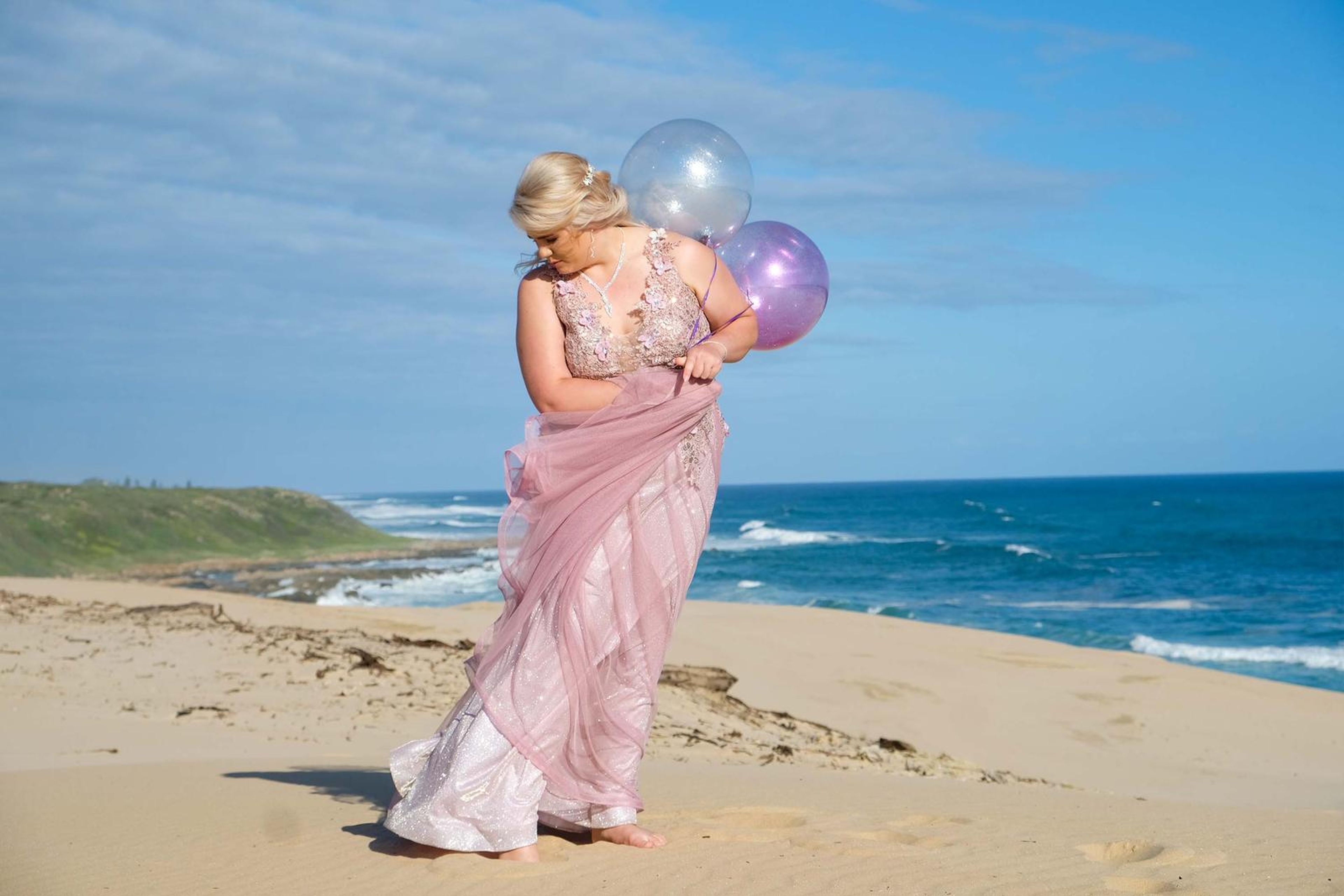 Holding balloons as the gown blows in the wind above the beach