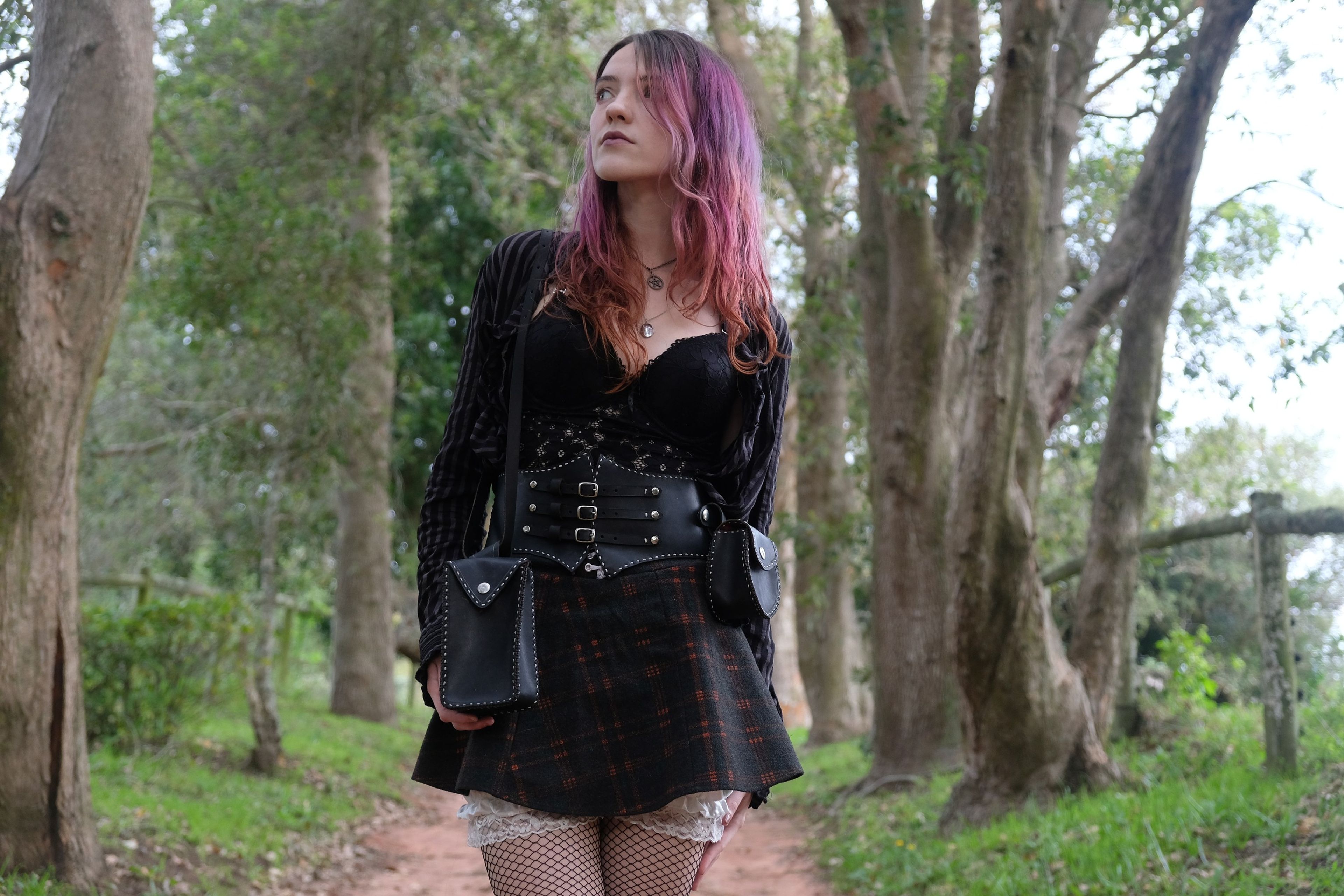 Woman in black standing on a path between trees, wearing a corset belt and a small bag