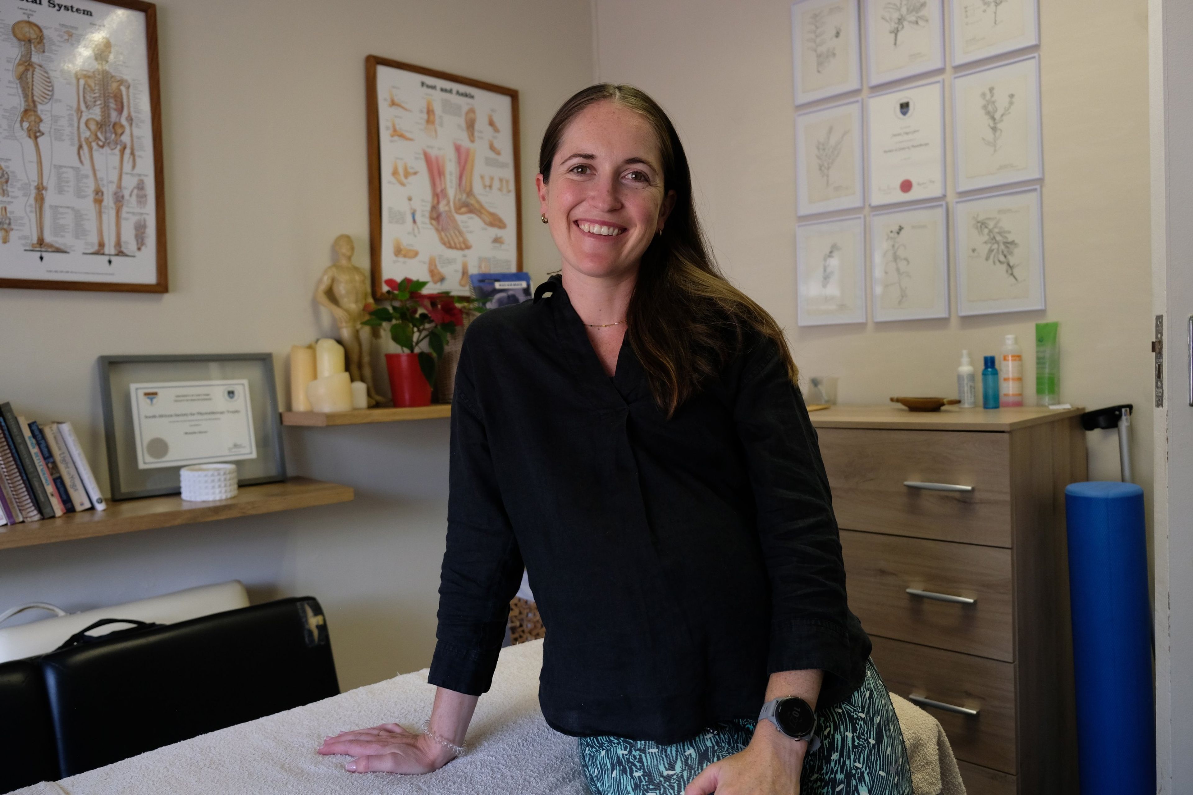 Team member in a black shirt leaning on a treatment bed and smiling, anatomy charts and framed certificates behind her