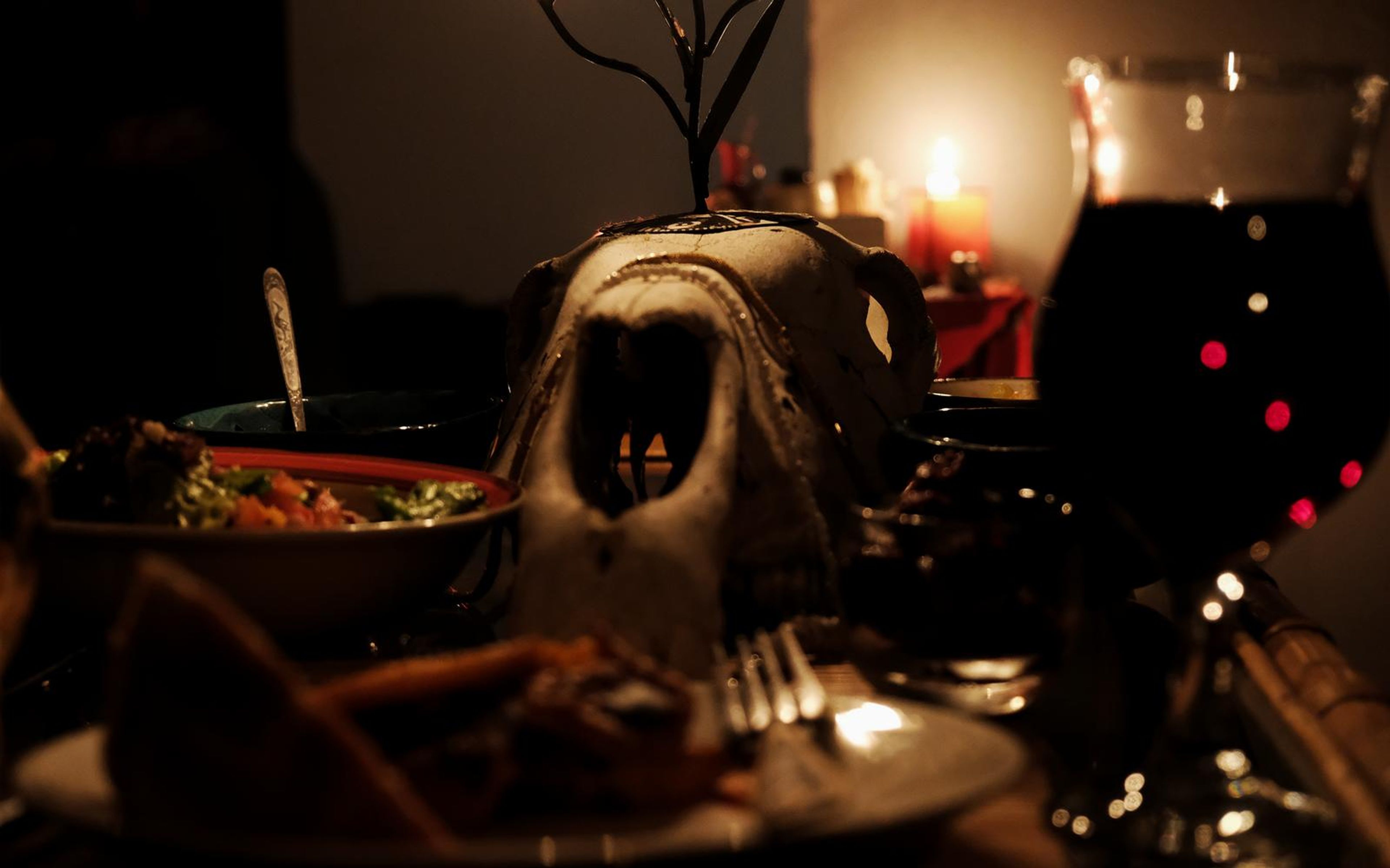Candlelit feast table with a pale skull, a bowl of food, a plate and a glass of red wine in warm low light